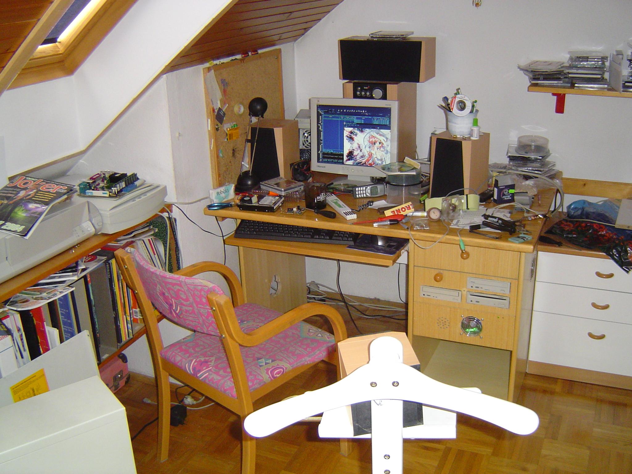 Very messy wooden desk, with LCD monitor in between two speakers with a subwoofer behind the monitor. On the left are some drawers, middle drawer having built-in 3 optical drives, and bottom drawer having a hole cut for a colourful 80mm fan, and some smaller holes for PC speaker. The computer is turned on.