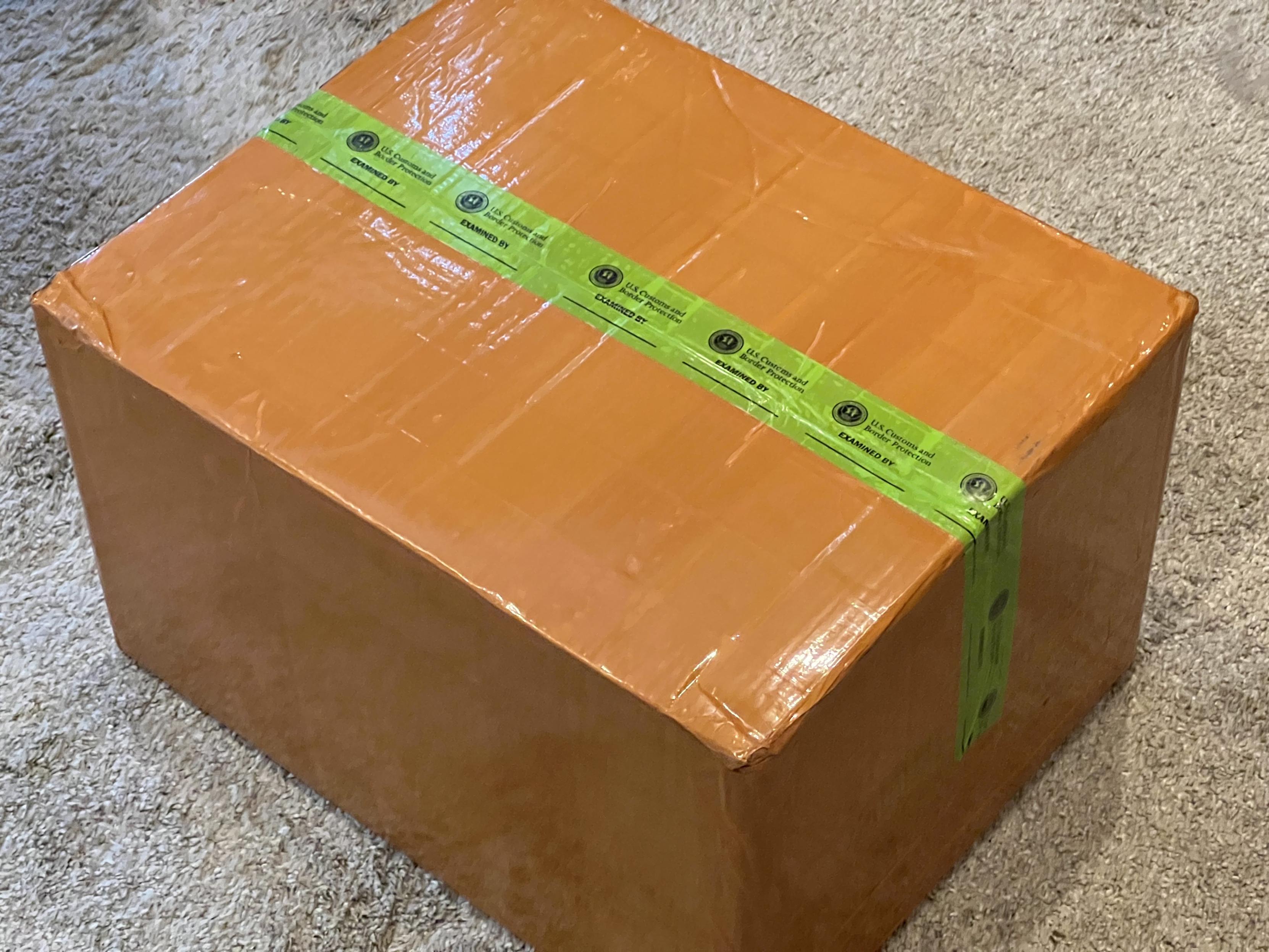 Photo of a cardboard box wrapped in orangish-brown tape, with the seam for the flaps having an extra green tape across it.