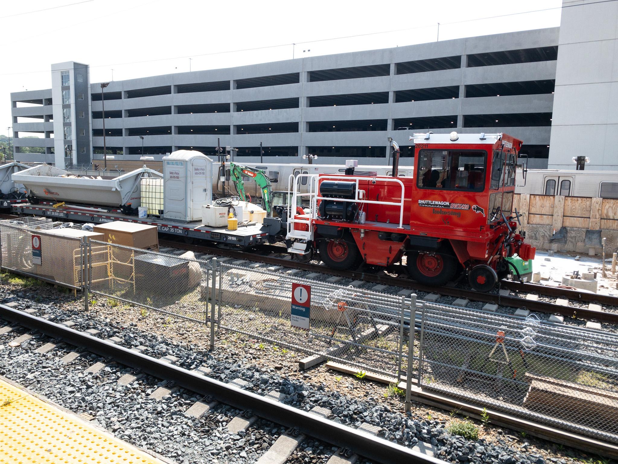 A small red diesel locomotive coupled to a flatcar that is carrying, among other things, a portable restroom. In the background is a parking garage.