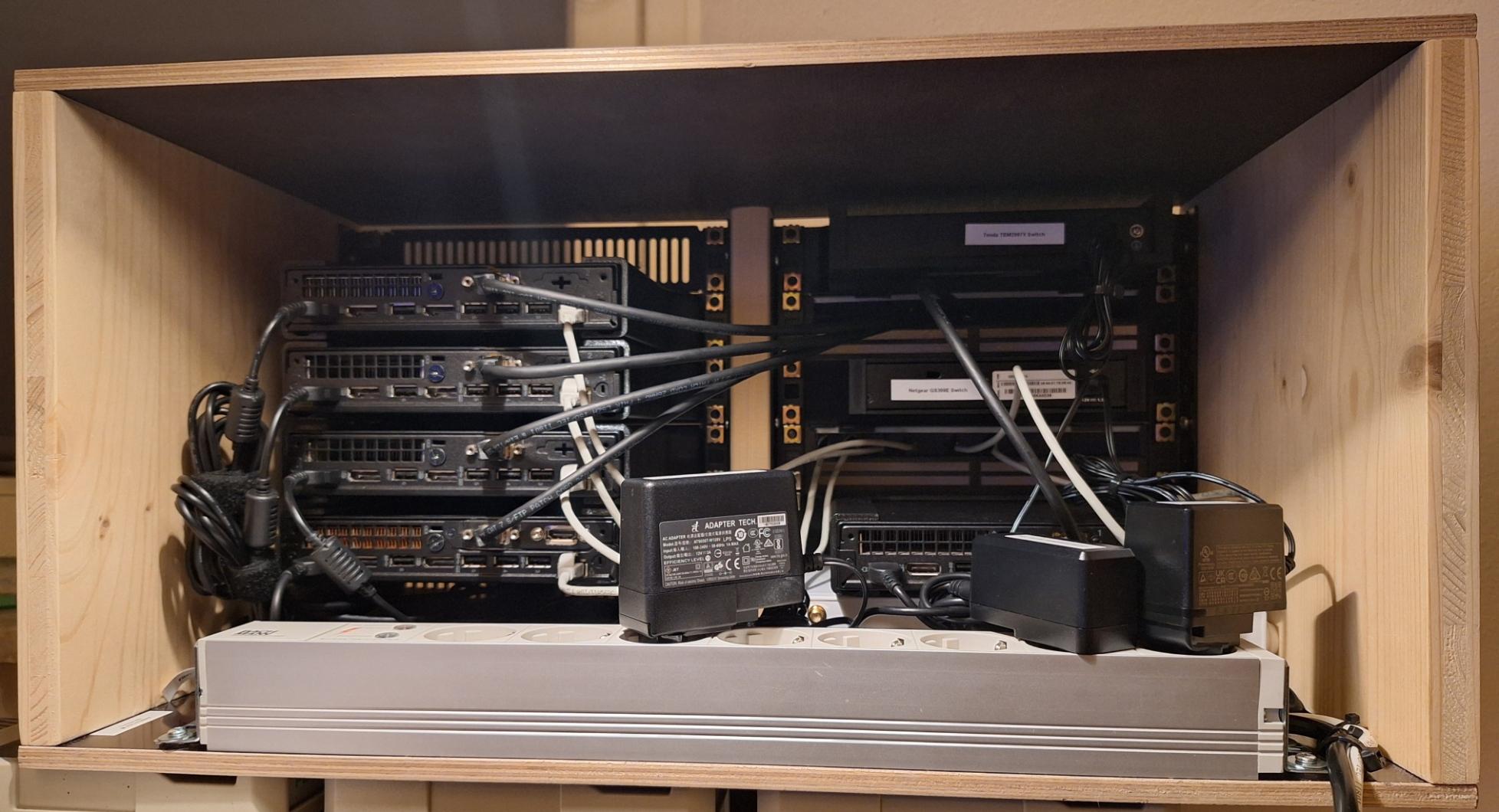 Ein Holzkasten, in dem zwei Netzwerkswitches, fünf Micro-PCs von Lenovo und eine Sophos-Firewall montiert sind. Ansicht von hinten ins Innere des Kastens.
In allen Geräten stecken Stromkabel. In den PCs stecken jeweils zwei Netzwerkkabel. Unten ist eine Mehrfach-Steckdosenleiste reingeschraubt, die aus silbernem Aluminium besteht. Darin stecken drei Netzteile.