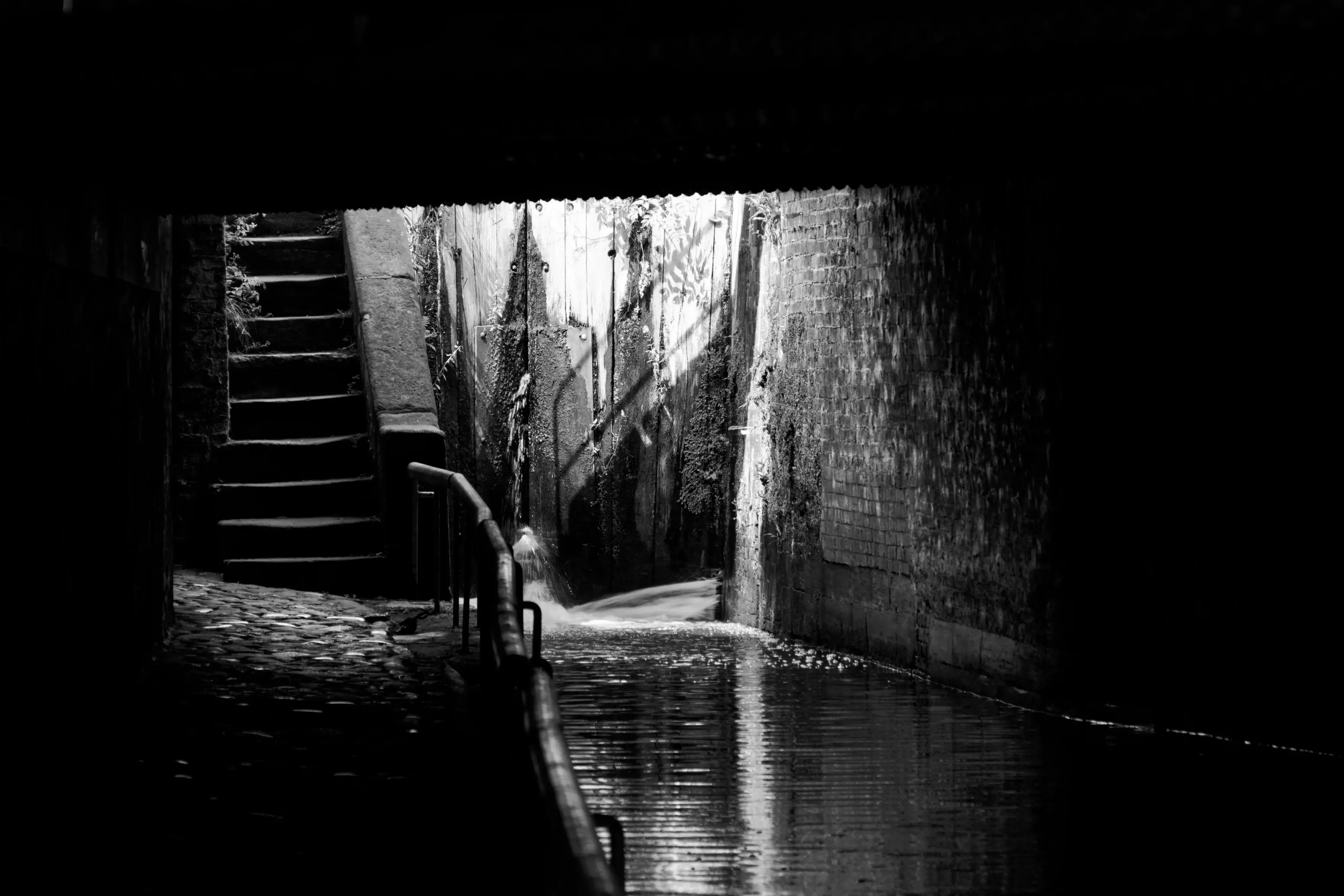 A black and white photograph captures a dramatic play of light and shadow inside a dark, covered brick tunnel.Bright sunlight streams from an opening above, sharply illuminating a stone staircase on the left that ascends out of view, a textured stone wall, and a wooden lock gate directly ahead. Water flows along a narrow canal channel on the right, creating a bright reflection on its surface. The left side features a dark path running alongside a metal handrail, with the foreground and sides shrouded in deep, heavy shadows.