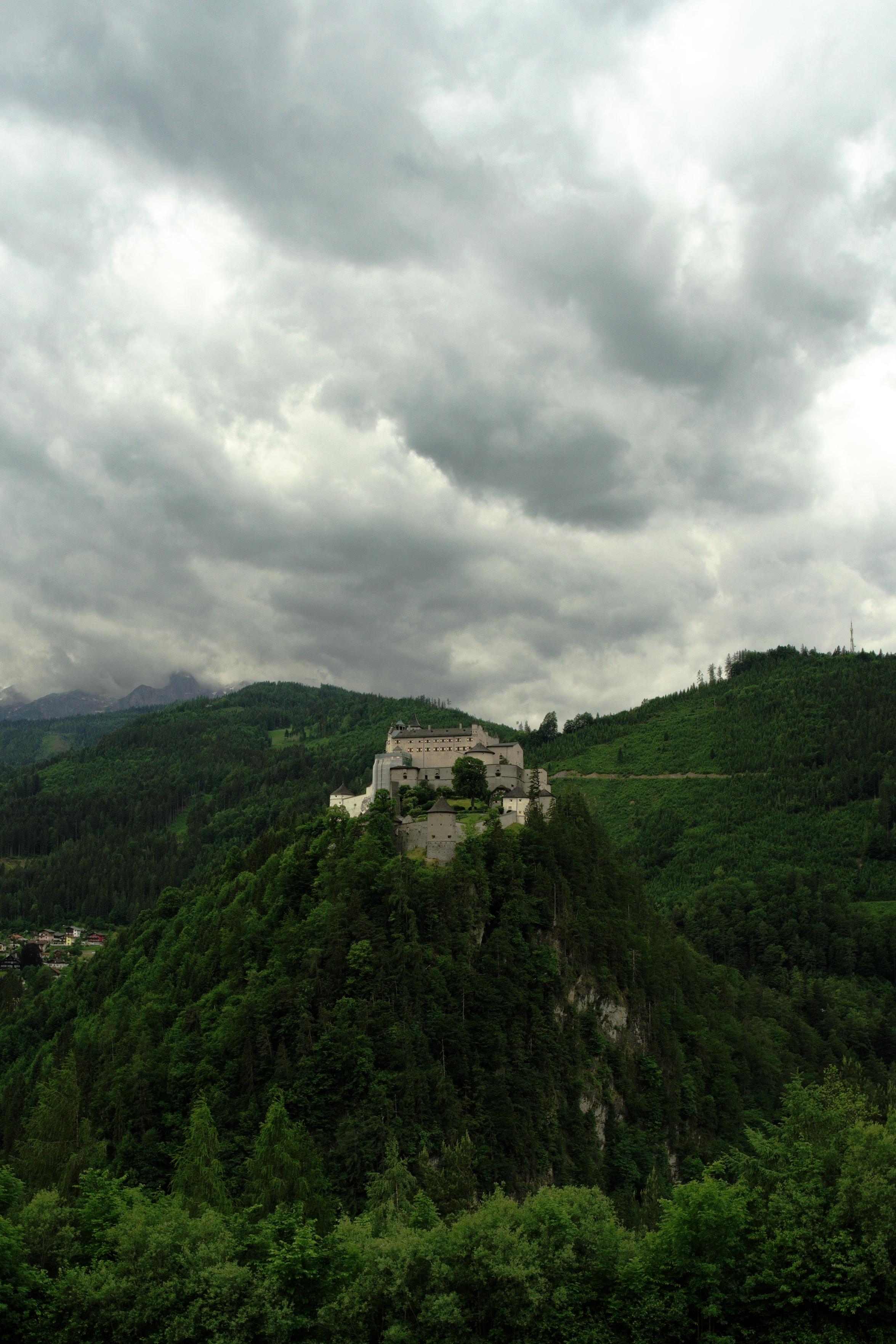 A photo of a castle on a hill with dramatic storm clouds hanging in the sky over the mountain in the background.