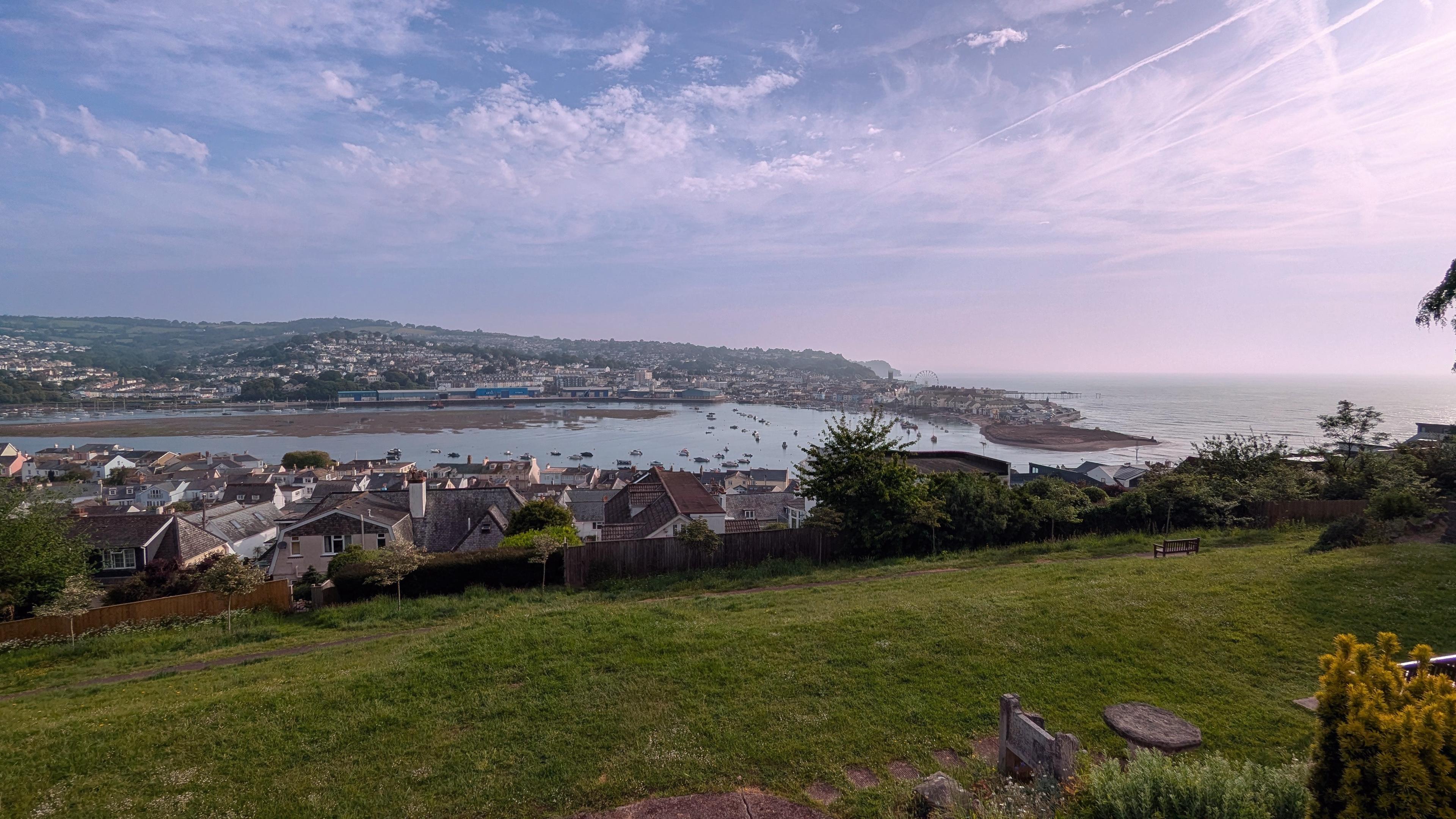 Starting with the near distance, this photograph has a green meadow sloping away to the rear of some houses in Shaldon, then onwards the estuary can be seen in between high and low tides with an area called the salty exposed, then onwards again is and the hell that rises above it giving way to a blue sky that is a little cloudy with the sun strongly towards the South or the right hand side of the picture.