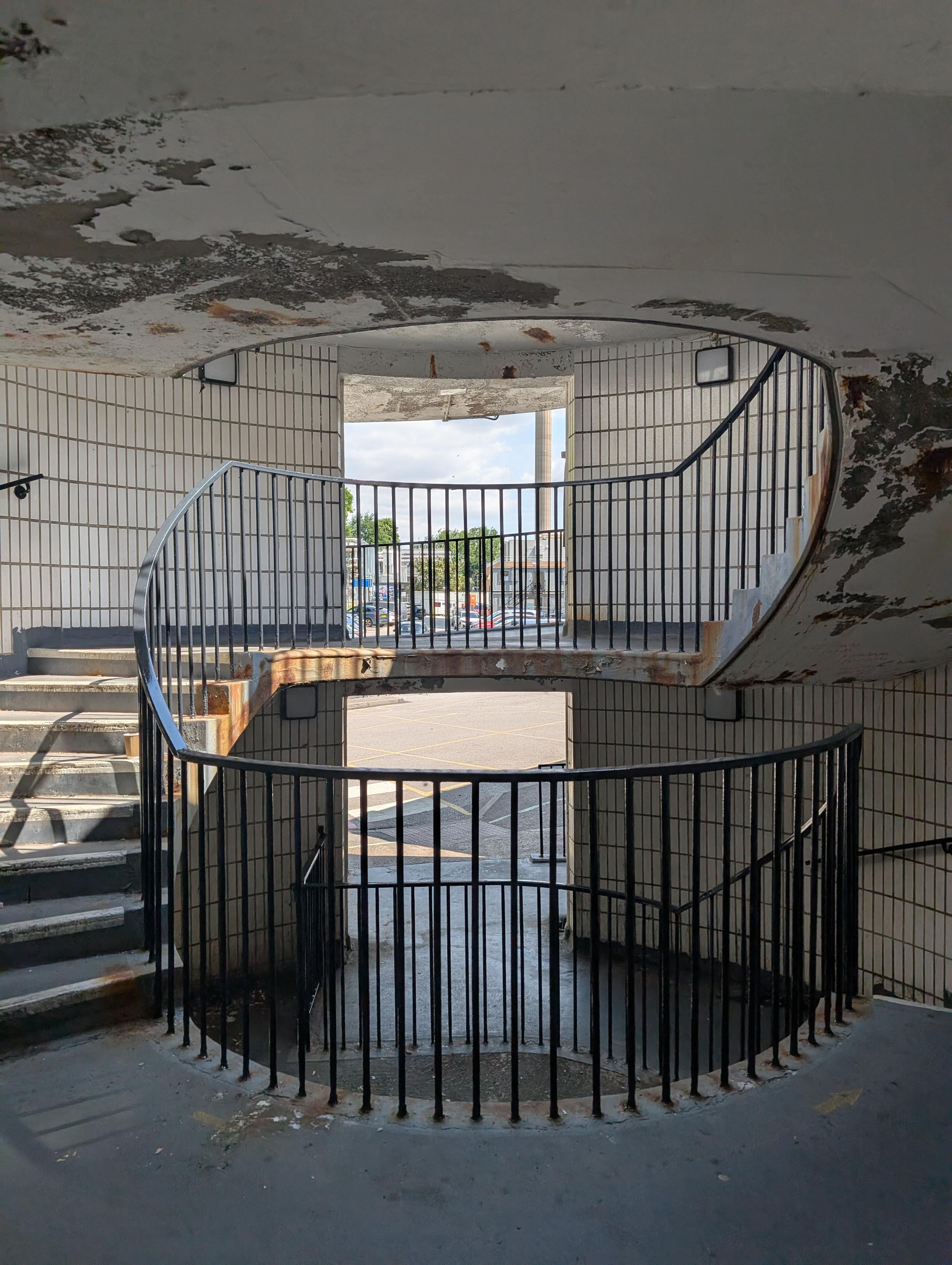 Concrete spiral staircase with black metal railings inside a white tiled stairwell. The ceiling shows peeling paint and weather damage. Central open portals look out onto an outdoor car park and bright blue sky