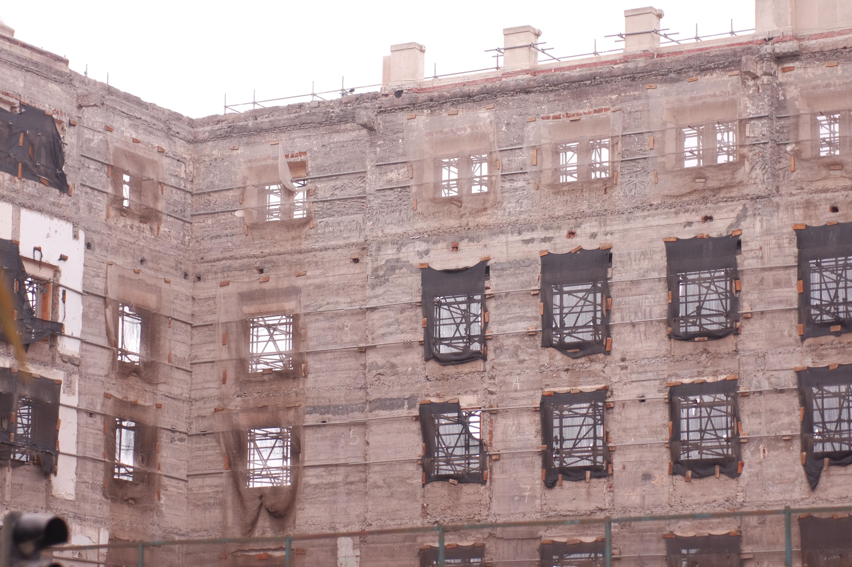 Exterior view of a multi-story concrete building under construction or renovation. Grid of rectangular window openings, some covered in black mesh netting, set against weathered, textured concrete walls with exposed scaffolding above.