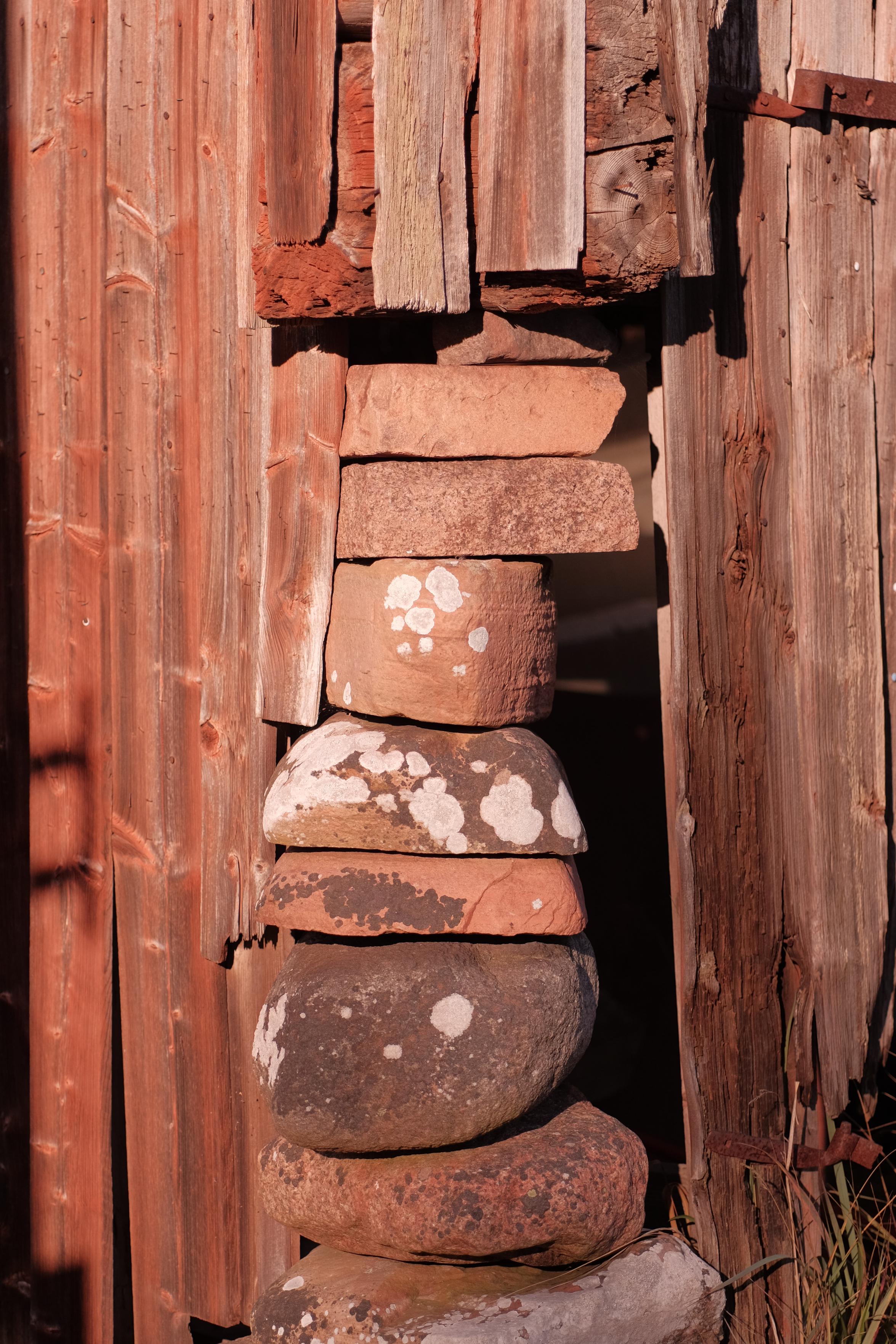 Vertical stack of flat, weathered stones stacked inside a gap between red-tinted, aged wooden planks. The stones are holding up the corner of an old agricultural building and they vary in color from light beige to dark gray, with white lichen spots visible on several surfaces.