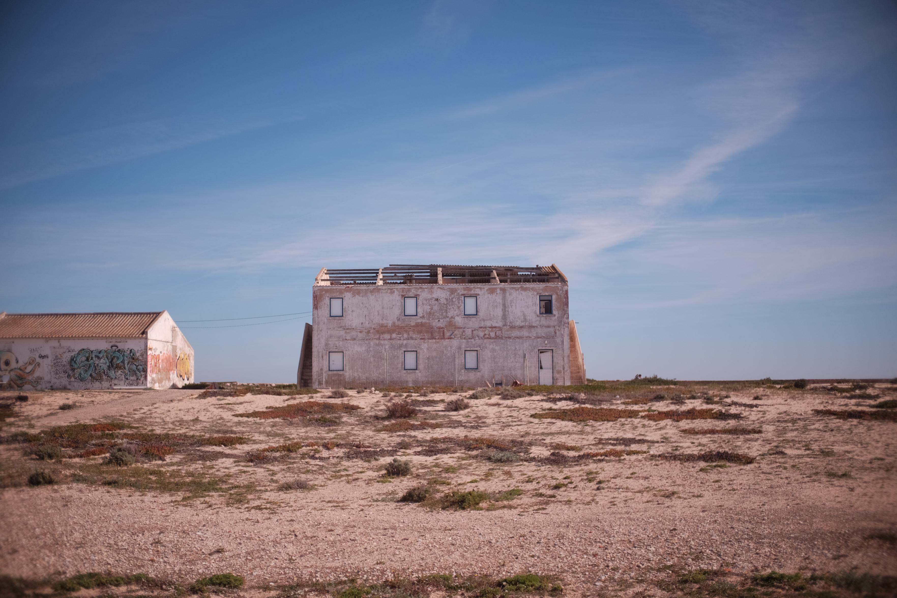 A two-story abandoned white building with an exposed, roofless top frame sitting in a flat, arid, sandy landscape under a clear blue sky. On the left, a smaller white structure features colourful graffiti on its side.