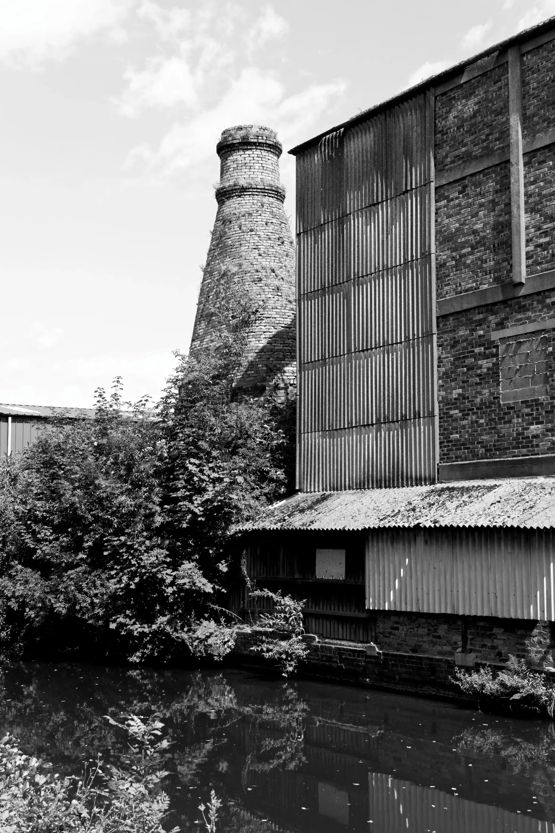 A black and white photograph captures a historical bottle kiln and industrial factory buildings situated alongside a calm canal.
On the left, the distinctive brick-built kiln tapers upward into a rounded chimney stack, partially obscured by dense leafy trees and bushes growing along the water's edge. To the right, a large multi-story factory structure features a combination of exposed brickwork and vertical corrugated metal siding. A low, single-story shed with a corrugated roof runs along the base of the main building, directly bordering the narrow canal. The dark, still water in the foreground reflects the industrial buildings and surrounding foliage under a lightly clouded sky.
