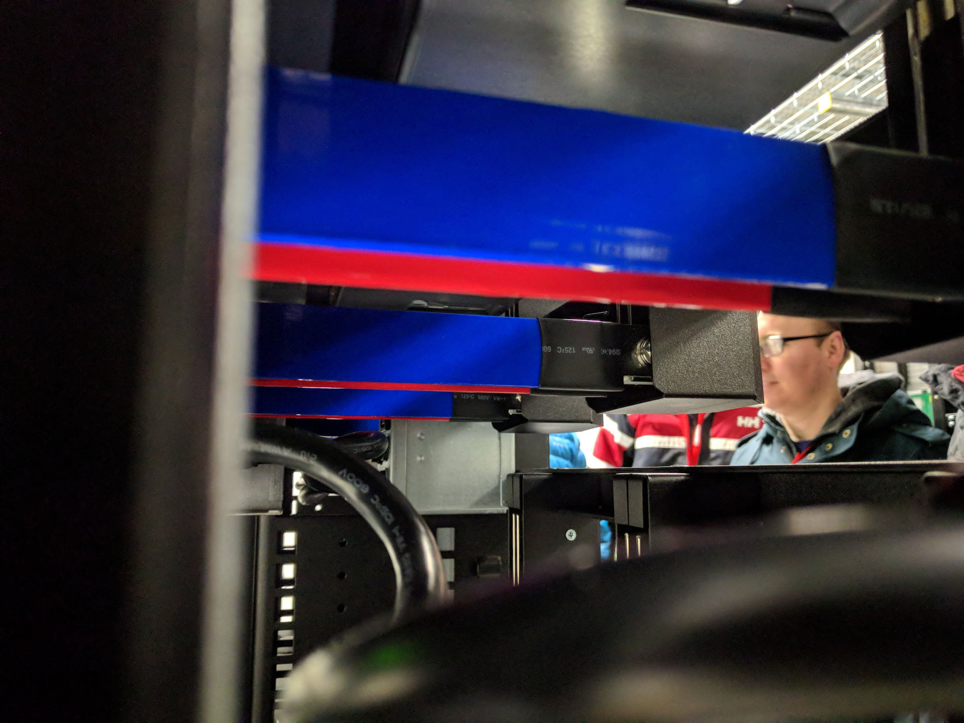 Die blauen und roten Stangen hier sind 12V Busbars für 12 V Gleichstrom mit echt vielen Ampere, die die zentralisierten Netzteile mit den Compute Units verbinden. Weil es weniger als 50 V sind ist keine Schutzisolation notwendig, nur funktionale Isolation.