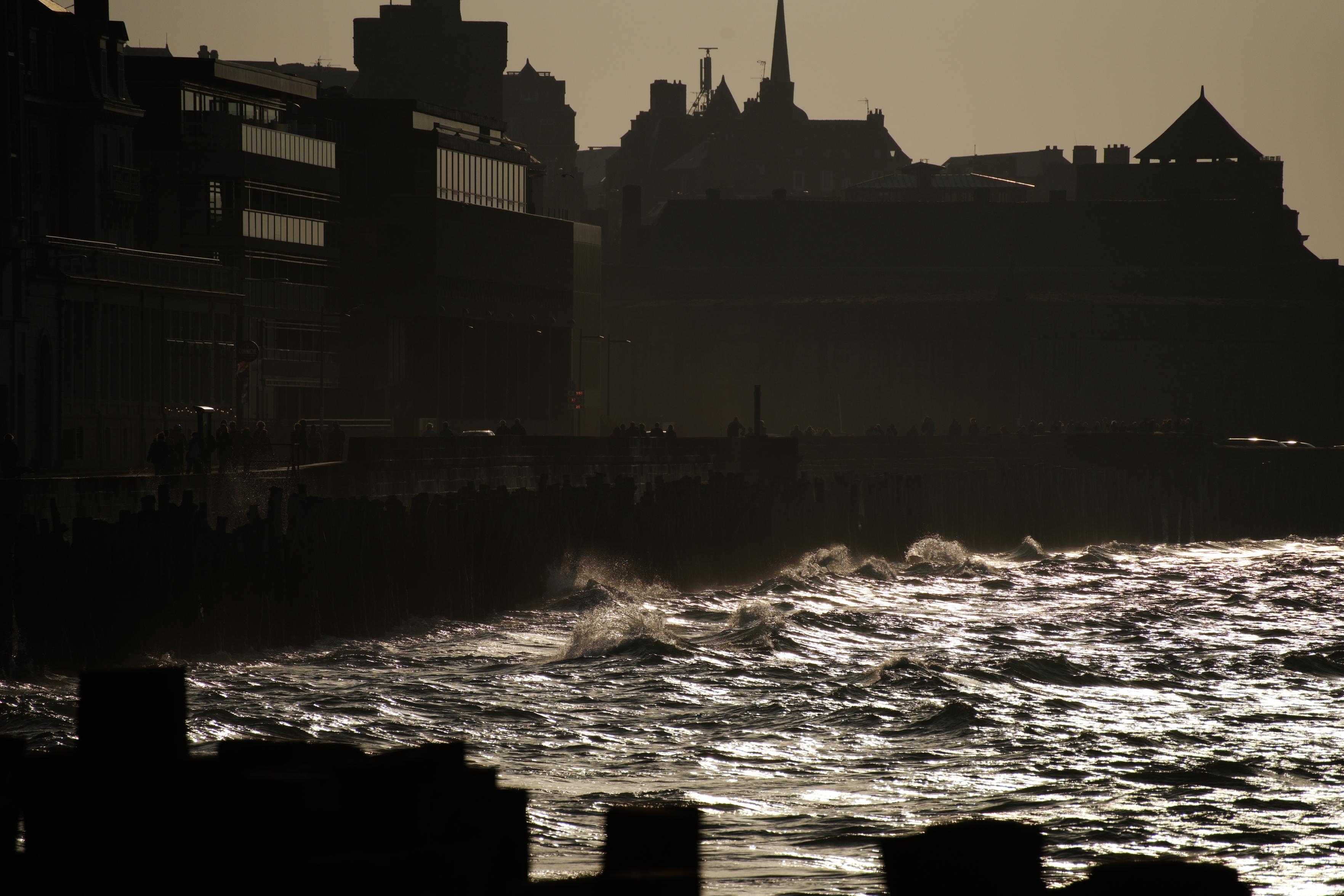 A photo of wavy water rolling against a seawall with buildings behind. The contrast is very high. The water is nearly white with reflections, the buildings dark and shadowy.