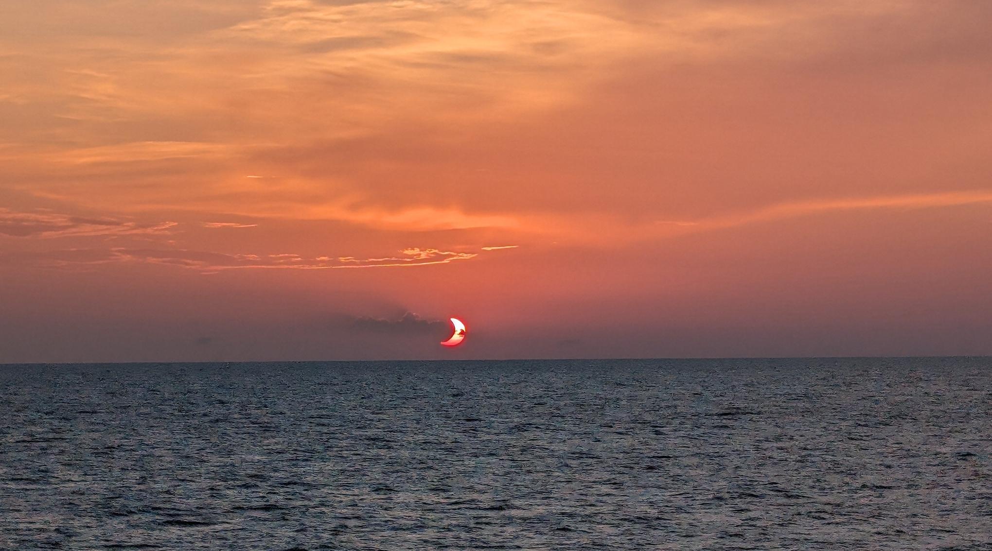a partial solar eclipse just above the horizon over the Mediterranean sea, with a cloud overlapping the moon's shadow on the sun -  off the coast of Mallorca