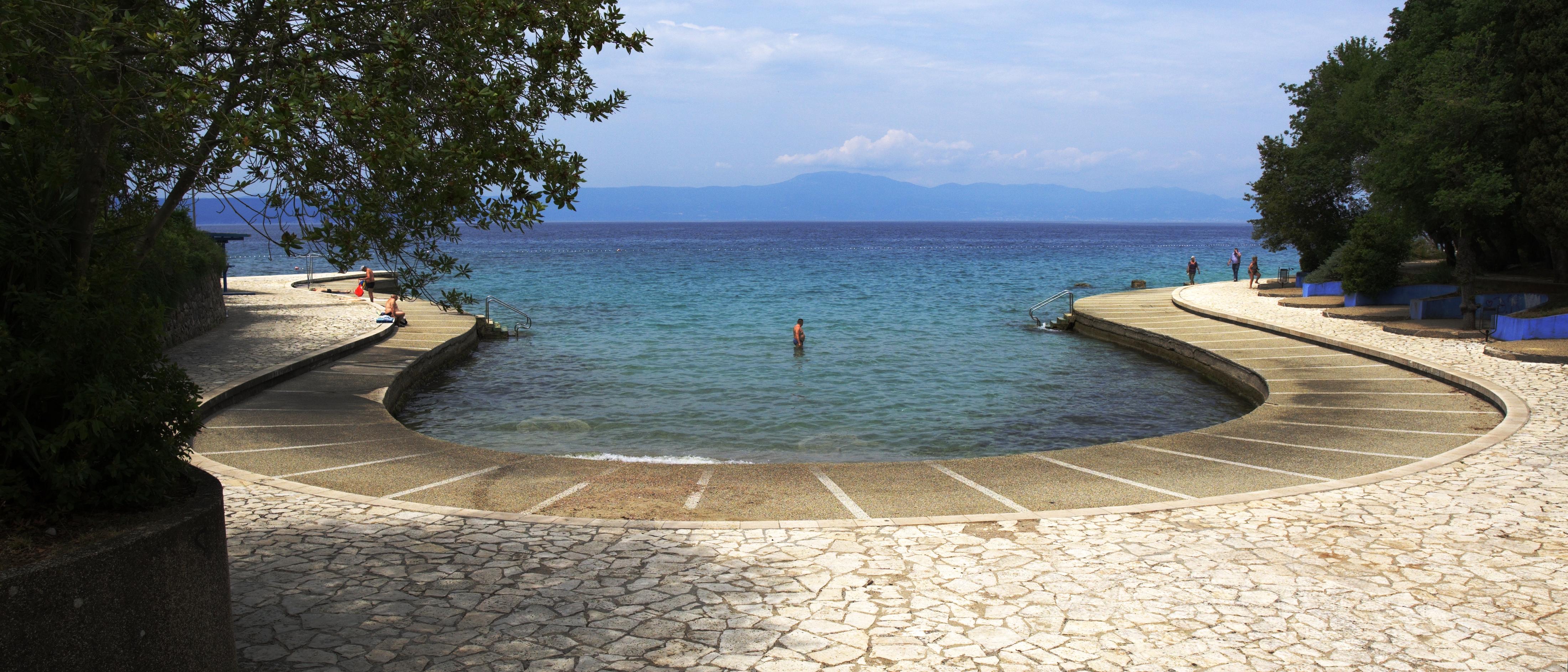 A photo of a circular pool bay by the sea with a single person standing right in the center. Lush trees on either side of the picture and a mountain range in the distance over the water.