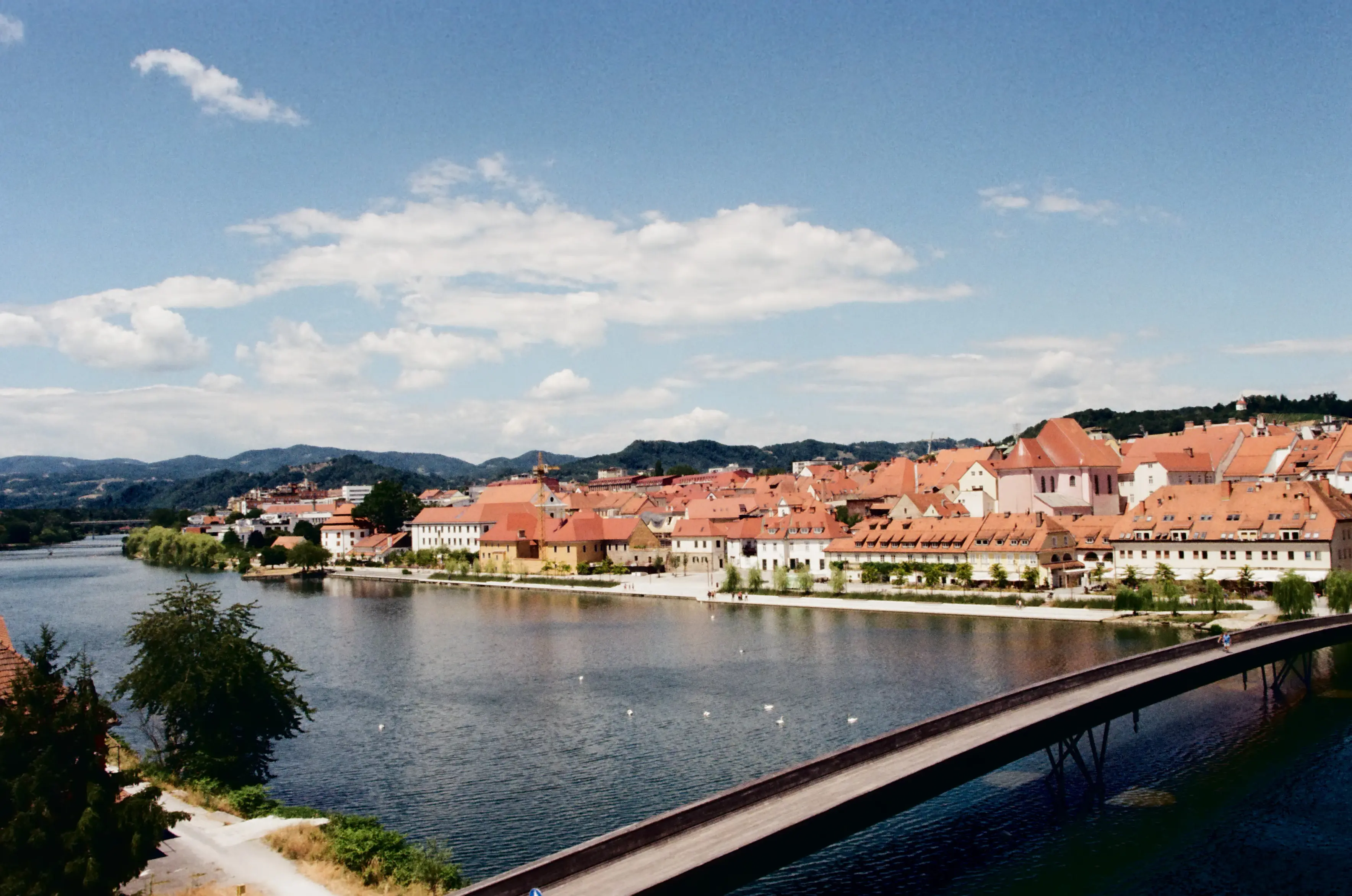 A wide view over the Drava River shows Maribor's old town on the far bank, its GORGEOUS red-tiled roofs by rolling green hills (or are they mountains?). A pedestrian bridge cuts diagonally across the foreground, with swans dotting the calm river below.