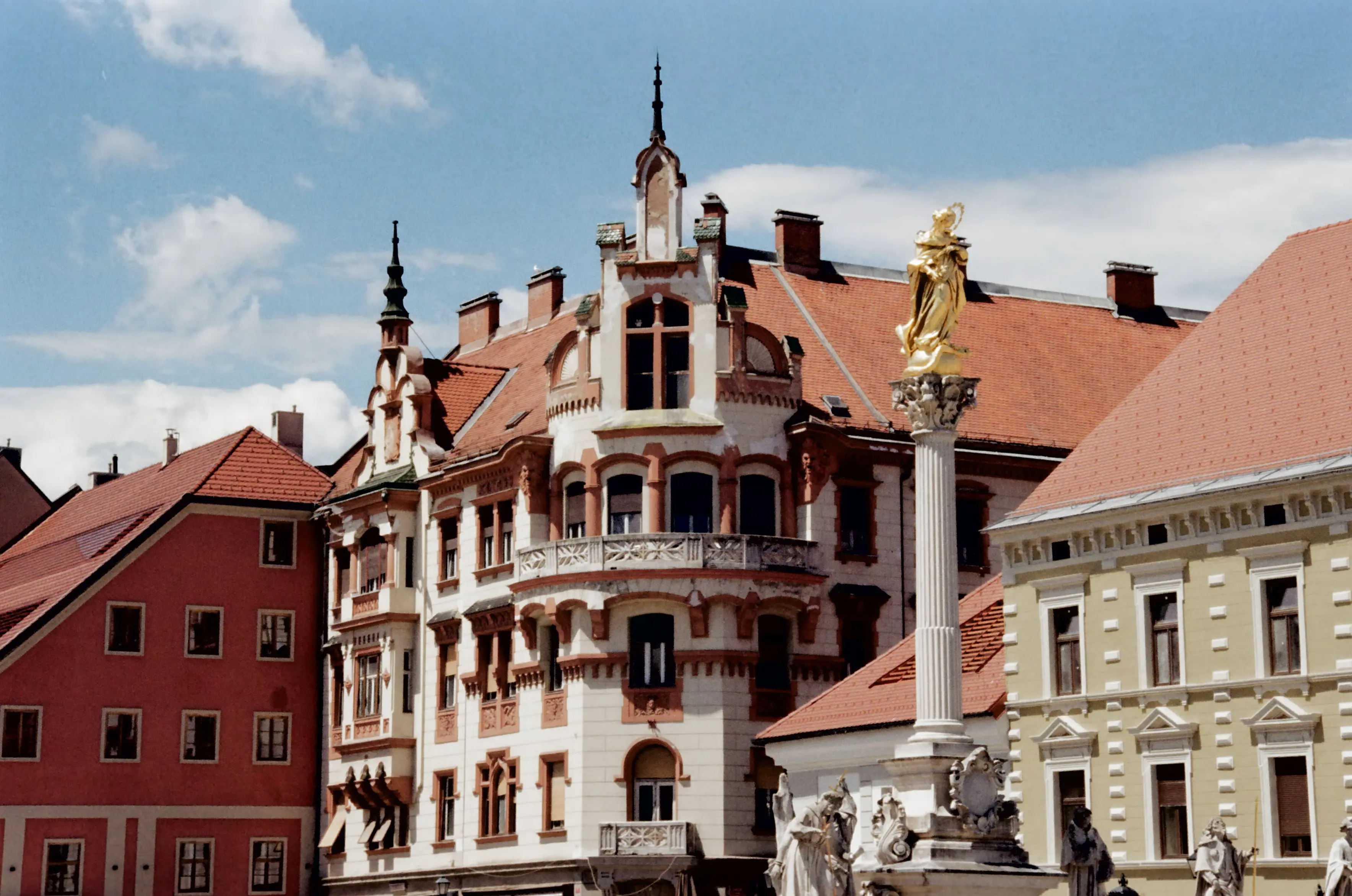 A gilded statue atop a column in Maribor's main square, standing before the white-red fancy facade of a corner building. Colorful row houses in red, cream, and white are in around this corner building, with warm film grain giving the whole image a nostalgic tone.