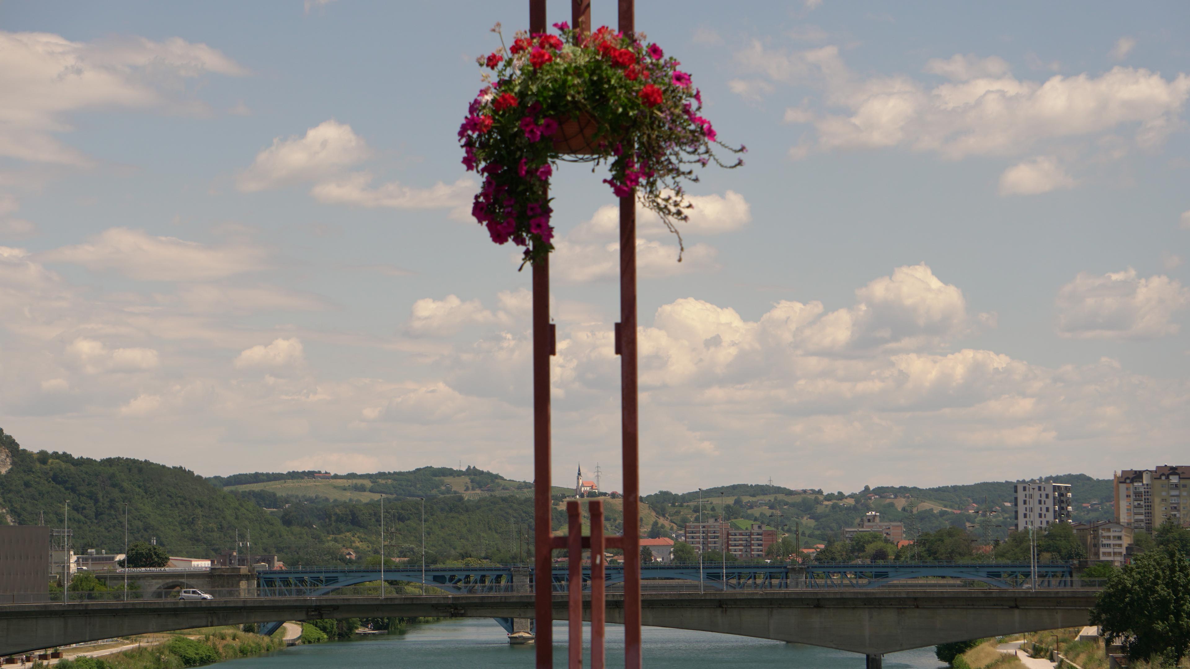 A flower basket frames a view of Maribor’s bridges, the Drava River, and surrounding green hills. Perfectly centered between the poles holding the basket is a church on the hills in the distance.