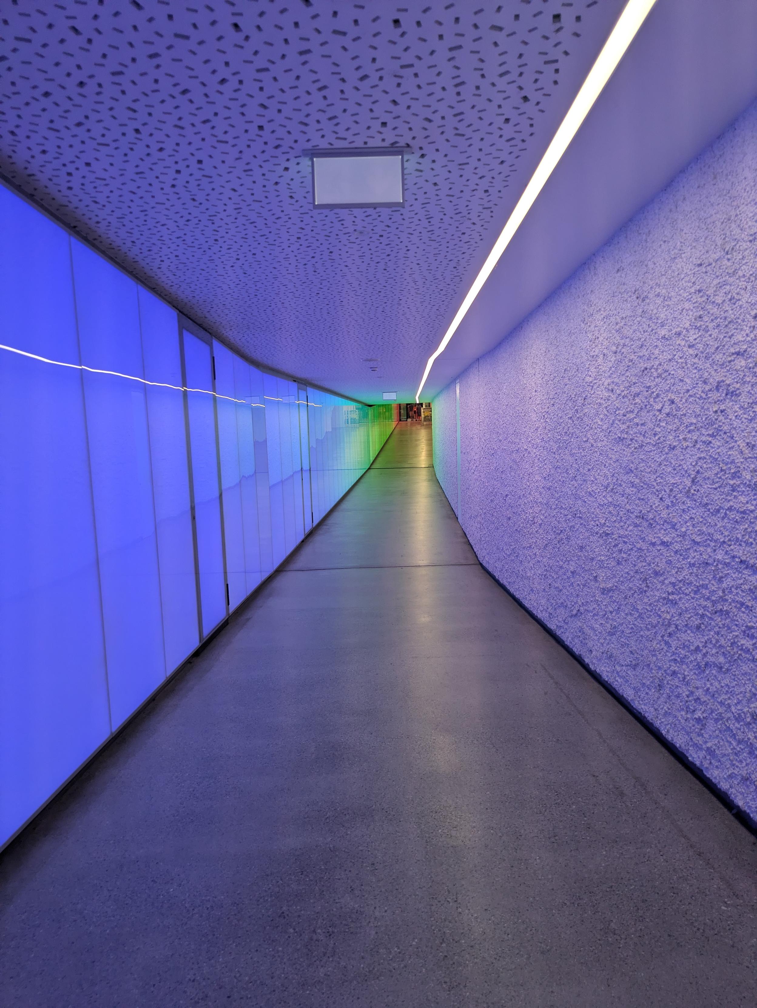 Taken in Thun, Switzerland. This modern looking tunnel leads from an inconspicuous entrance in the old town, via a car park and many, many stairs, to the castle above the town. Well worth a visit if you are nearby.

In terms of composition, the tunnel is curved. Vanishing point upper center frame. The light emanating from the left hand wall is pale blue and purple but green in the distance. My guess is that the cross section would be three by three meters. The ceiling has a contiguous strip of white light a few centimeters wide.