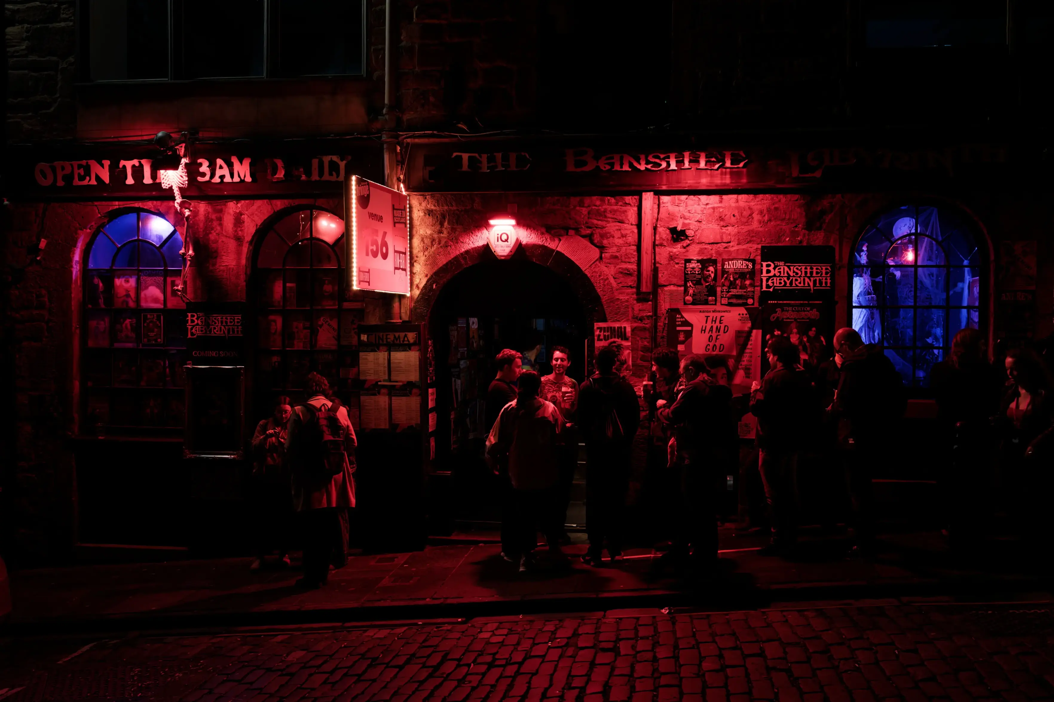AI Mode conversation: write alt textYou sent: 1 image and said: write alt textwrite alt textA nighttime, eye-level wide shot captures the illuminated stone storefront of The Banshee Labyrinth pub in Edinburgh, Scotland.
The pub features a dark grey stone facade with a large arched central entryway where a small group of people are gathered. To the left and right of the entrance are two large arched windows with blue interior lighting. Posters, flyers, and event signs are affixed to the stone walls and windows, including a prominent white rectangular sign with the number "156" in red. Above the entrance, a dimly lit horizontal sign reads "THE BANSHEE," and another sign to the far left states "OPEN TIL 3AM DAILY" in red neon lettering. The entire scene is bathed in a dramatic crimson and purple glow from the pub's exterior and interior lighting, which reflects onto the dark cobblestone street in the foreground.