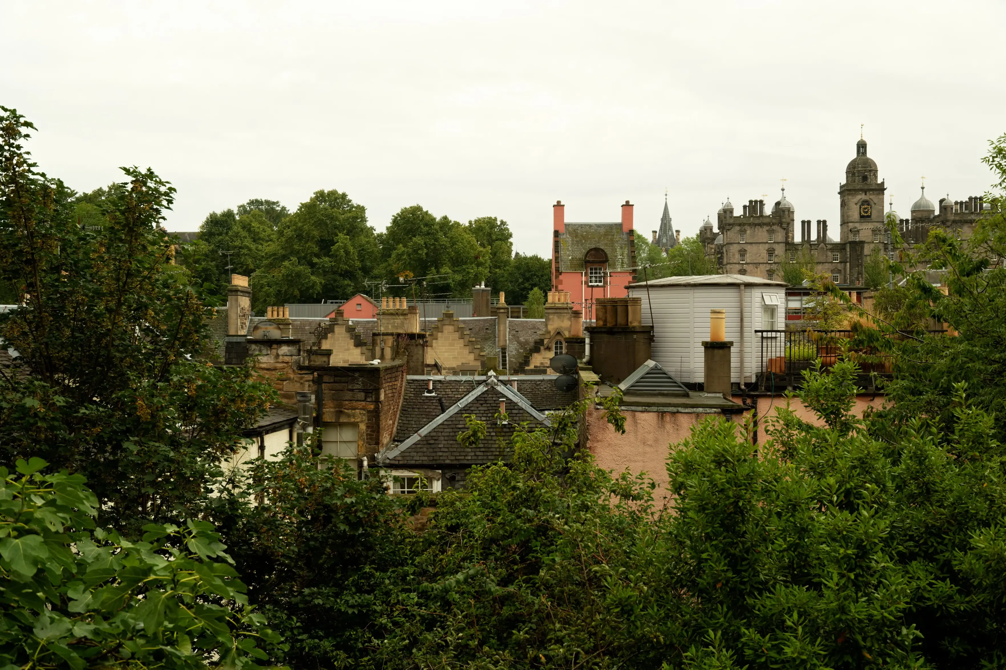An elevated, wide view overlooking historic stone rooftops, stepped gables, and chimneys framed by lush green trees in the foreground. In the mid-ground stands a distinctive pink, steep-roofed building, while historic stone architecture with turrets and a clock tower rises in the background under an overcast, hazy sky.