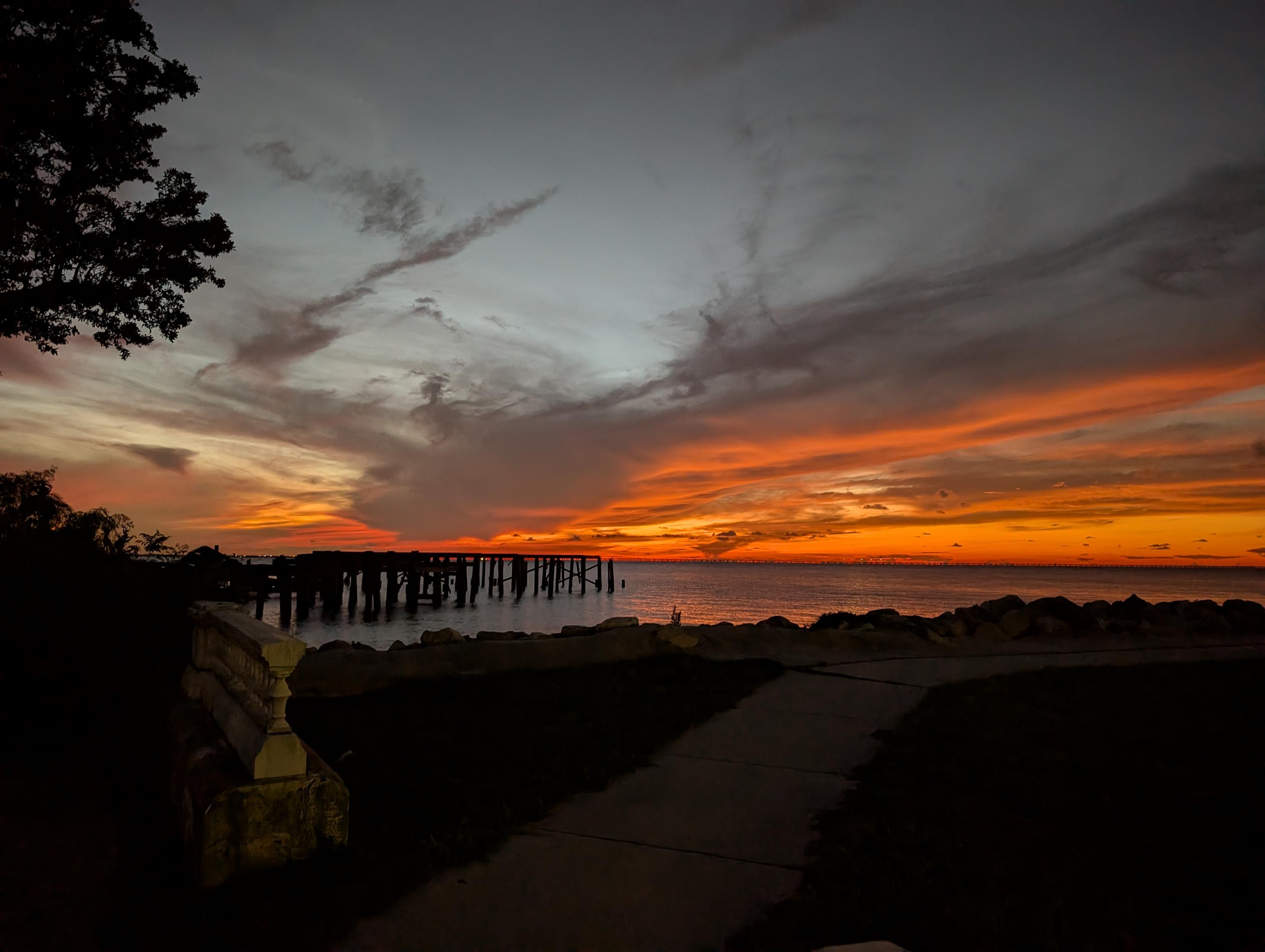 A very red sunset over the water. A path extends off to the right. Old pilings extend out into the water.