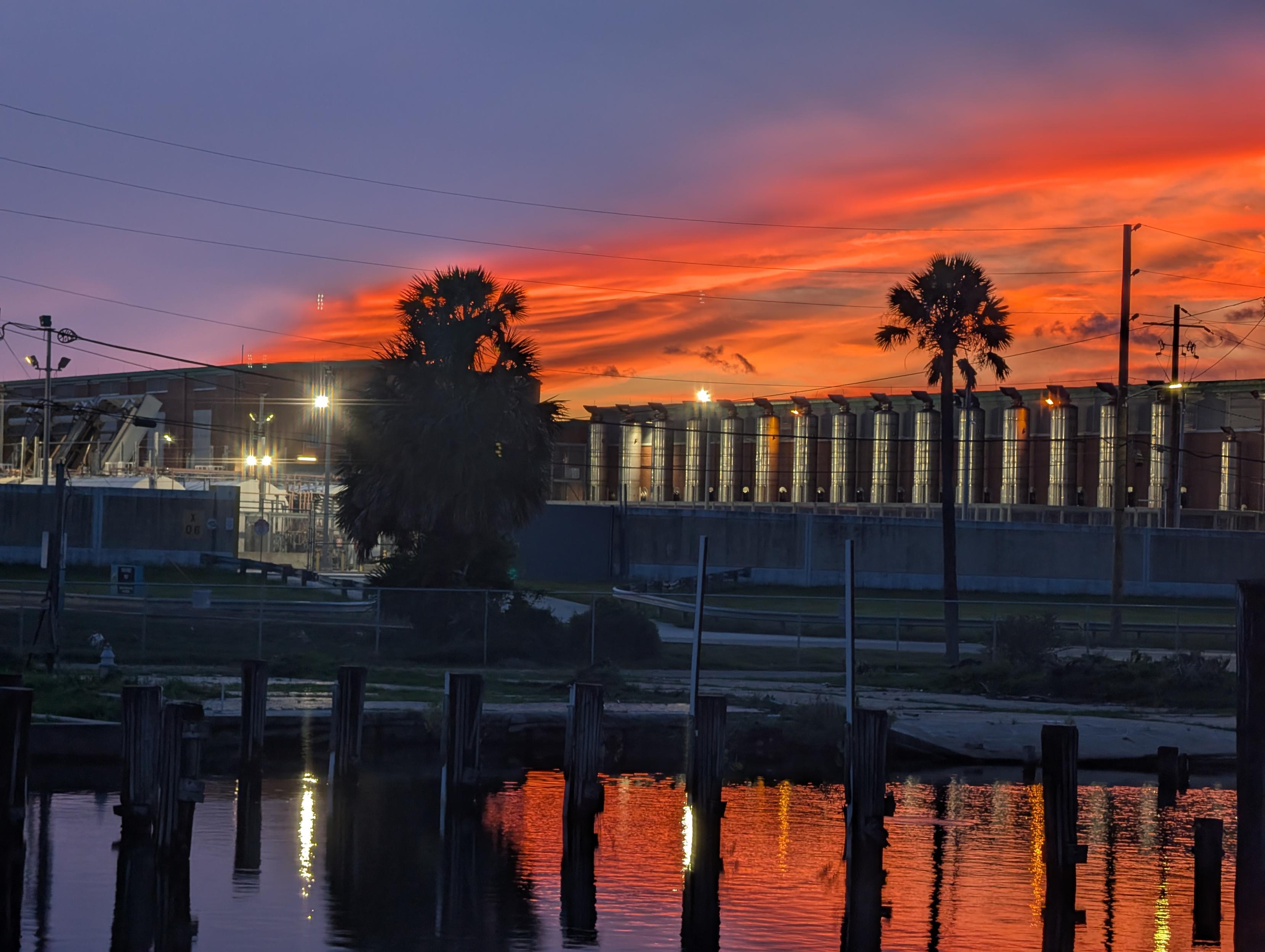 Harsh lights illuminate a pumping station. The red sky above is reflected in the water below.