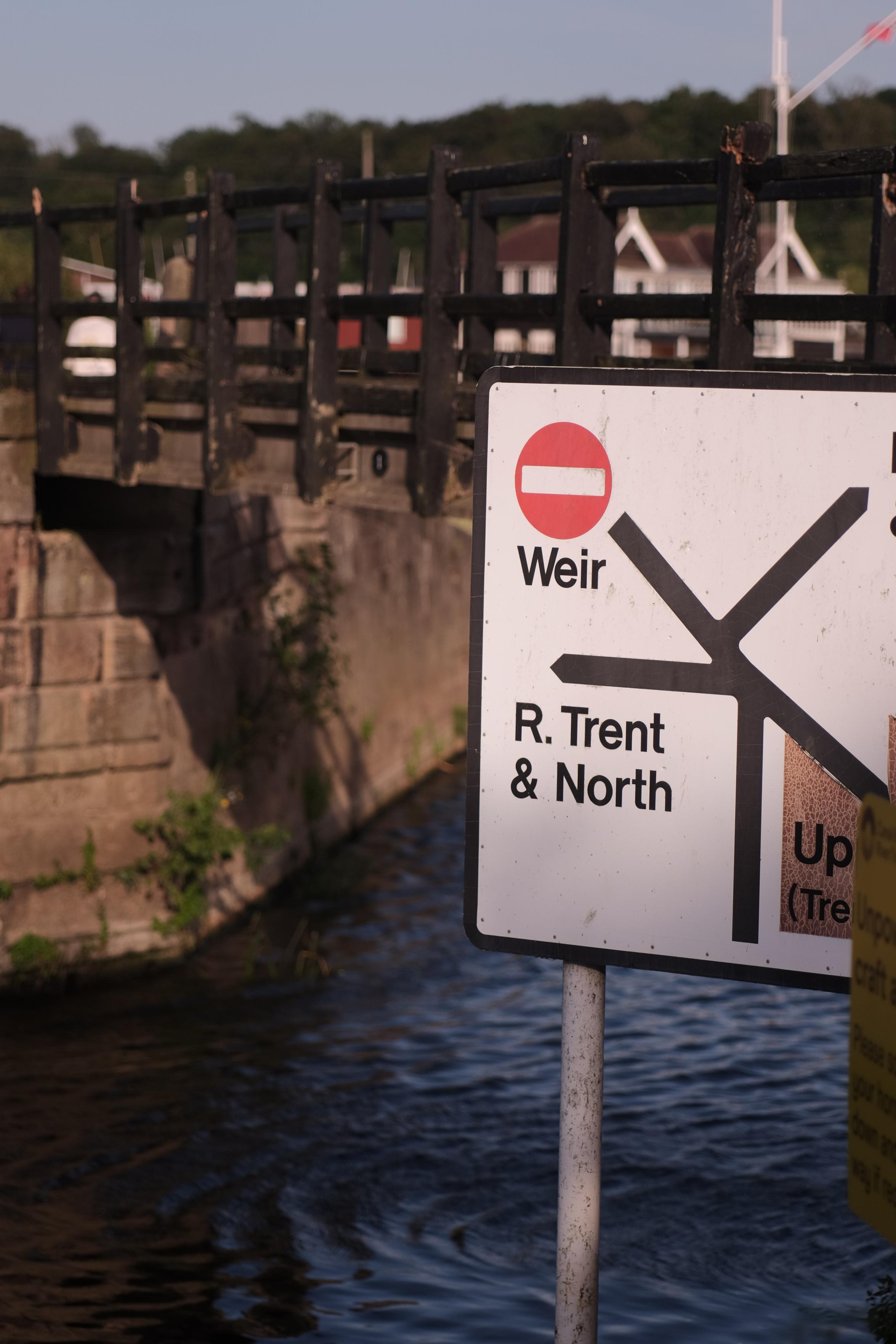 Navigational signpost beside a dark waterway with a stone and wood bridge in the background. The white sign features black directional lines. A line pointing upper-left has a red no-entry symbol and reads "Weir". A line pointing left reads "R. Trent & North". A line pointing right reads "Up (Trent)".