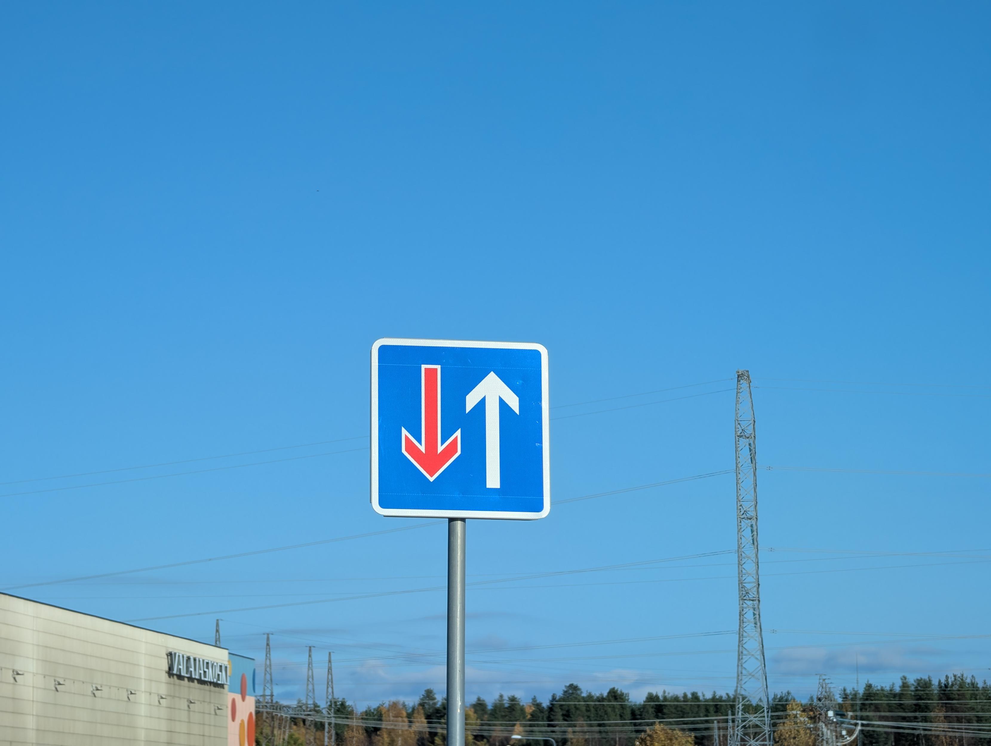 Square blue traffic sign against a clear blue sky, with a commercial building, trees, and an electrical tower in the lower background. The sign features a white border and displays a red arrow pointing down on the left alongside a white arrow pointing up on the right.