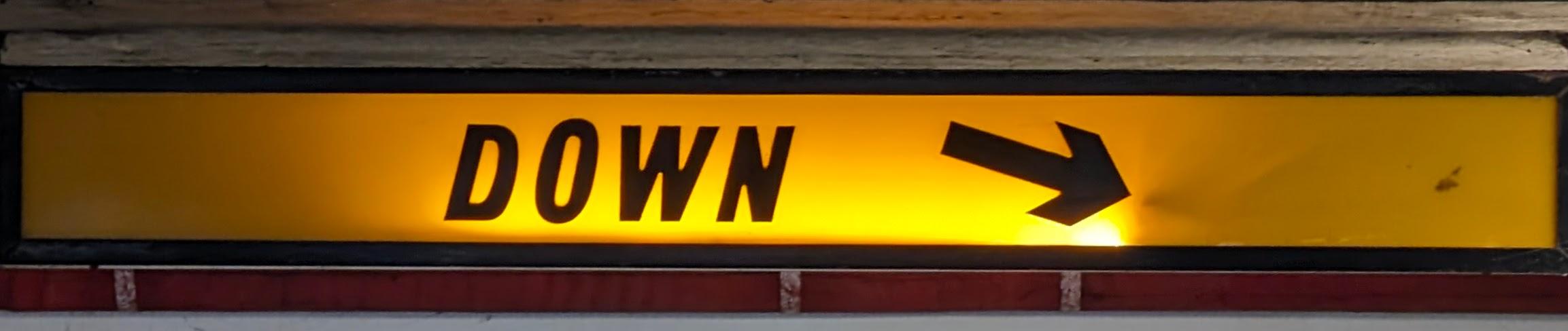 Illuminated yellow rectangular sign with a black frame, mounted between a light-colored ledge and a row of dark red tiles. The sign displays the text "DOWN" in black block letters, followed by a thick black arrow pointing to the right.