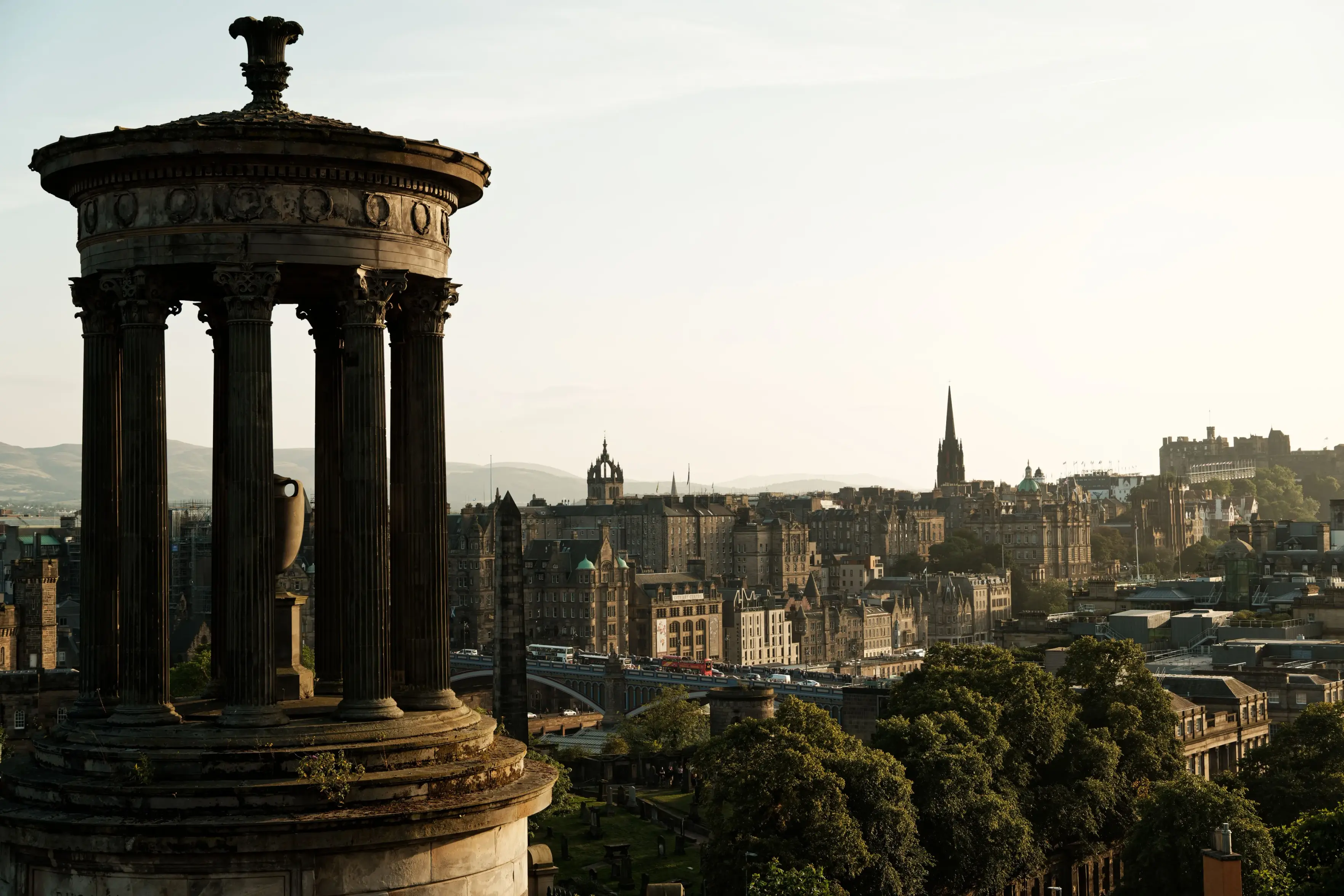 A high-angle, wide shot captures the Dugald Stewart Monument overlooking the historic city skyline of Edinburgh, Scotland, under a clear, warm sky.
In the left foreground, the weathered stone monument features a circular temple structure with Corinthian columns and an ornate urn at its center. Beyond it, the dense urban landscape stretches across the frame, showcasing classic Gothic and Victorian architecture with numerous spires, stone buildings, and a bridge bustling with traffic. Lush green treetops fill the lower right foreground, while Edinburgh Castle stands prominently on a rocky hill in the far right background, all bathed in soft, golden sunlight.