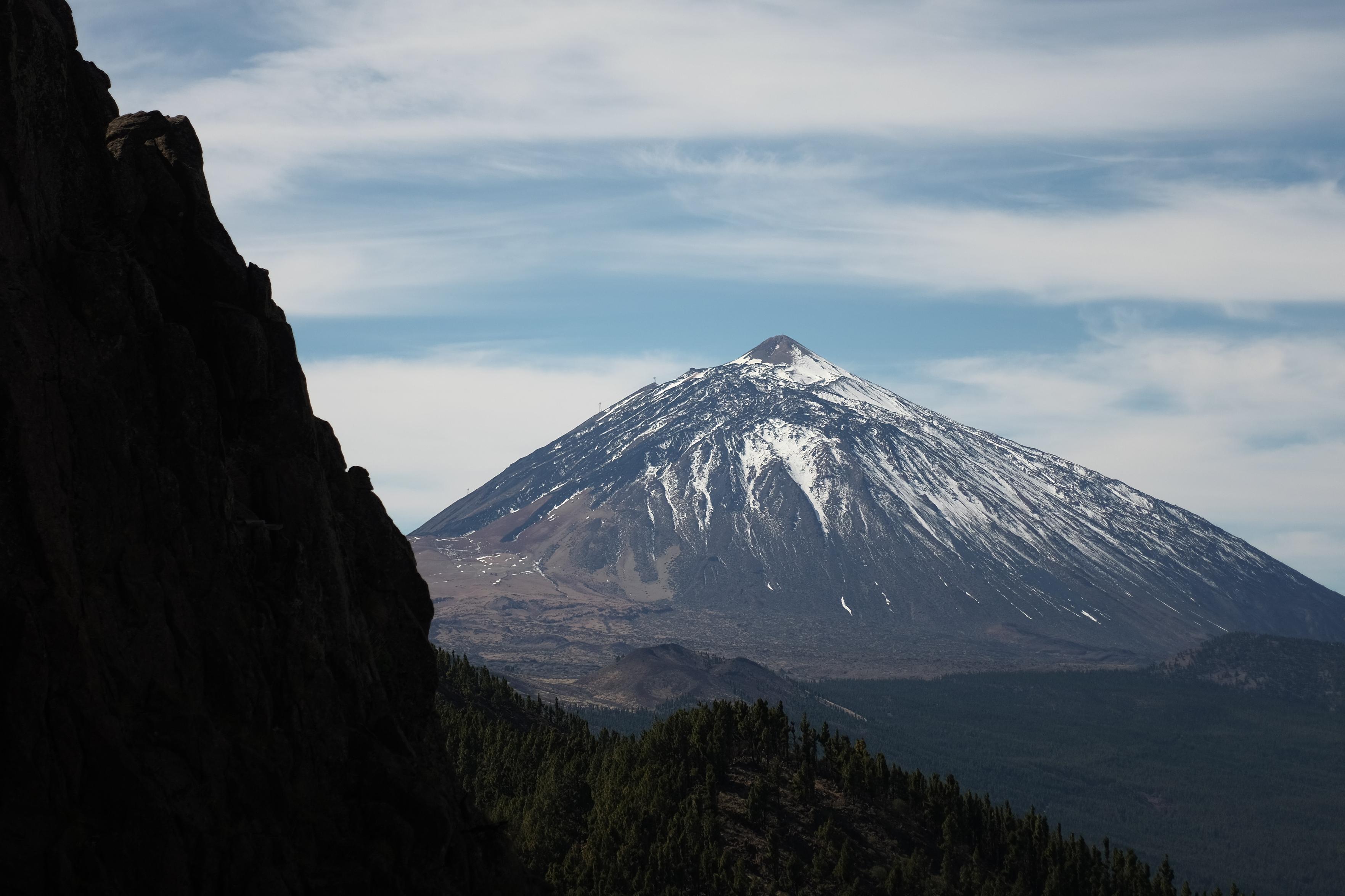 A snow-dusted, triangular volcanic peak rises under a blue sky with thin, wispy white clouds. A dark, jagged rock wall frames the left foreground in shadow, while dense green pine trees cover the sloping ridges below the mountain. The slopes of the peak display alternating stripes of white snow and dark rock.