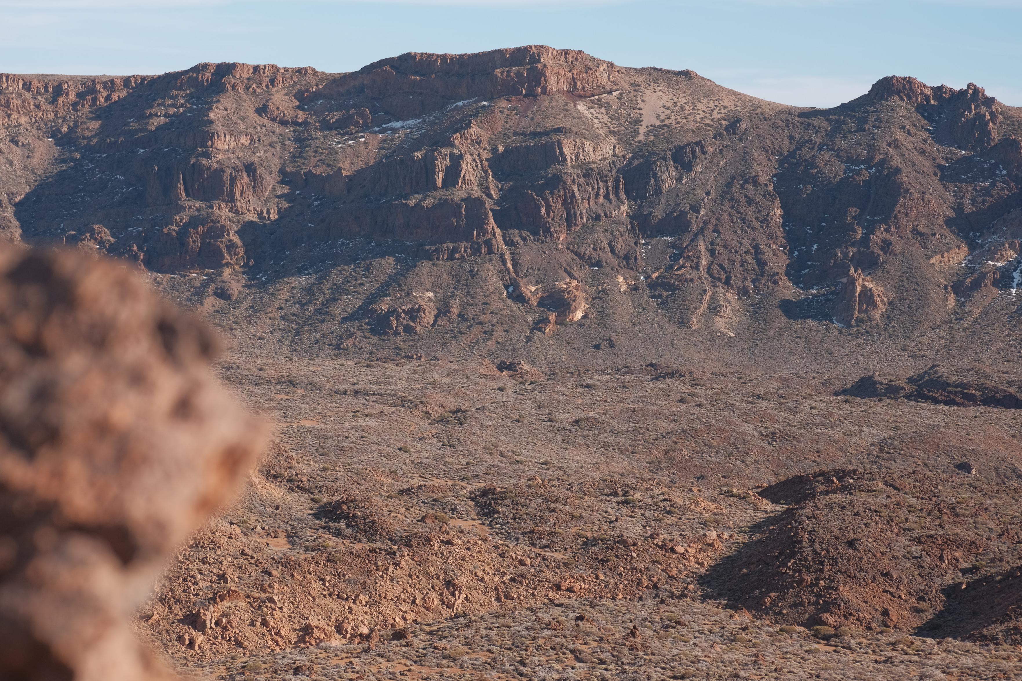 Arid, rugged mountain landscape under a pale blue sky. A broad volcanic plateau with steep, rocky cliffs and layered brown ridges spans the background. The foreground and valley floor consist of rolling terrain covered in dry, brown and tan brush, with an out-of-focus rock formation visible in the lower left corner.