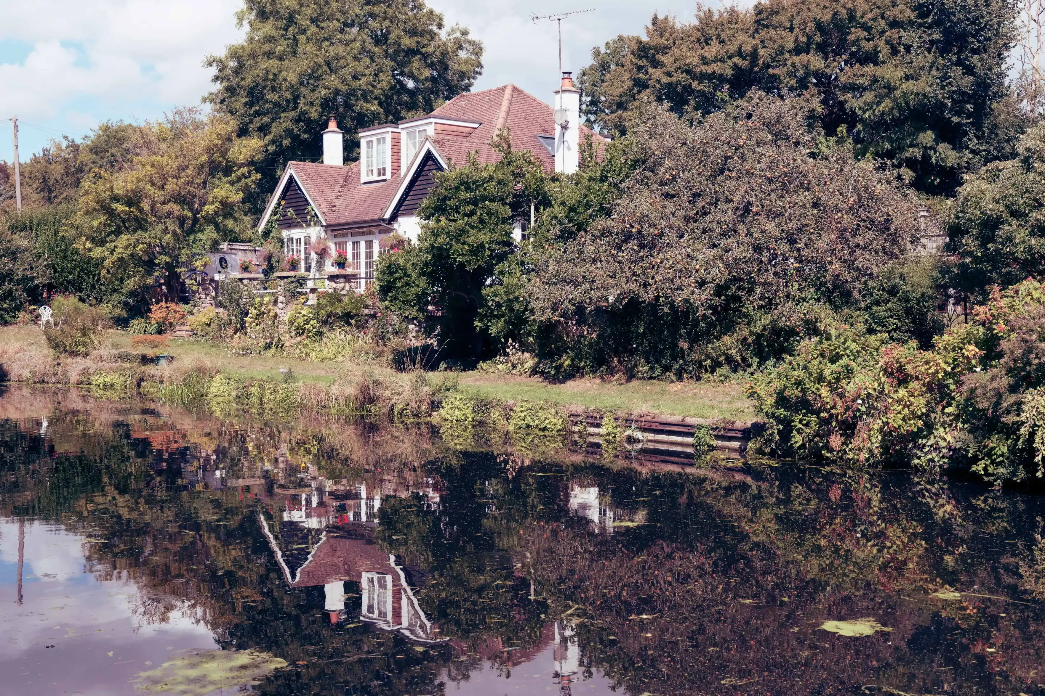 A cottage with a reddish-brown tiled roof and white walls nestled amongst lush green trees and dense foliage, casting a clear reflection in the still water of a canal in the foreground under a partly cloudy sky.