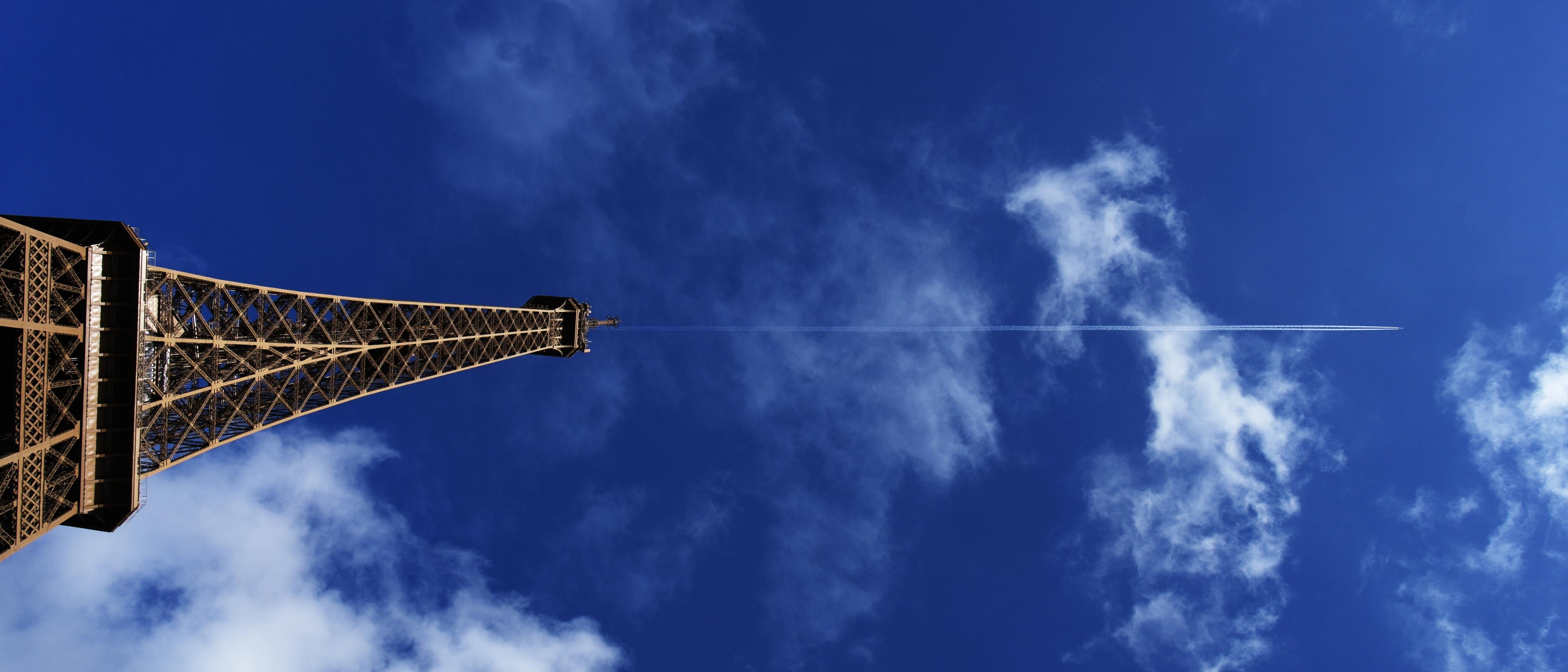 A picture of the top part of the Eiffel Tower on the left side of the frame with a plane flying overhead, the contrail nearly aligned with the tip, going right.