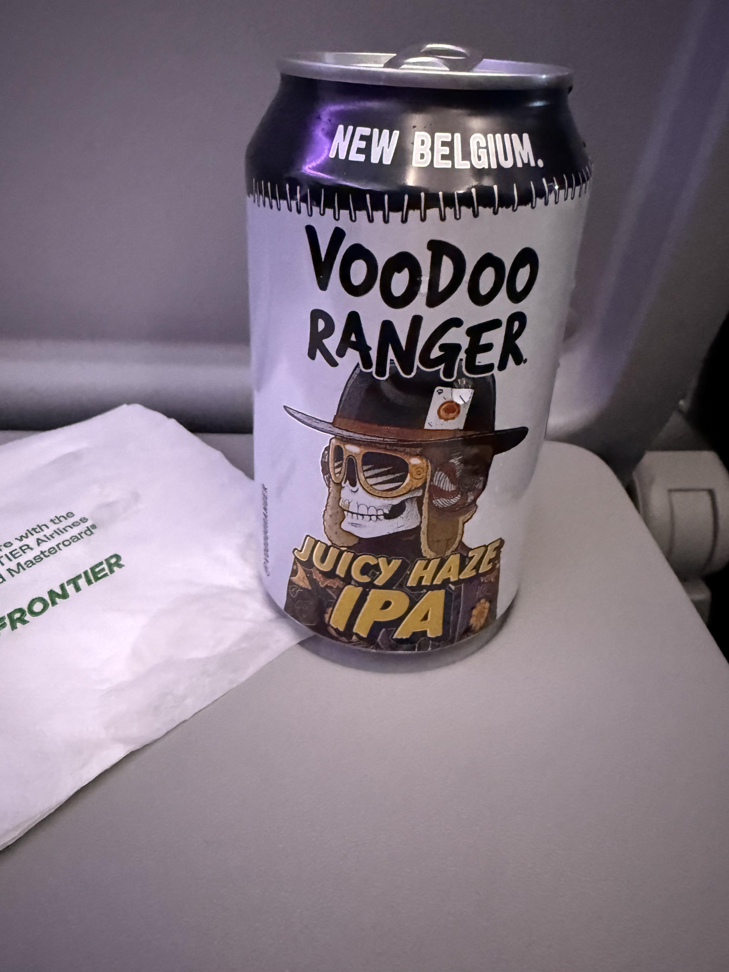 A white can with a black top band sits on an airplane tray table. A sunglass wearing skull with a swanky hat is pictured on rhe can