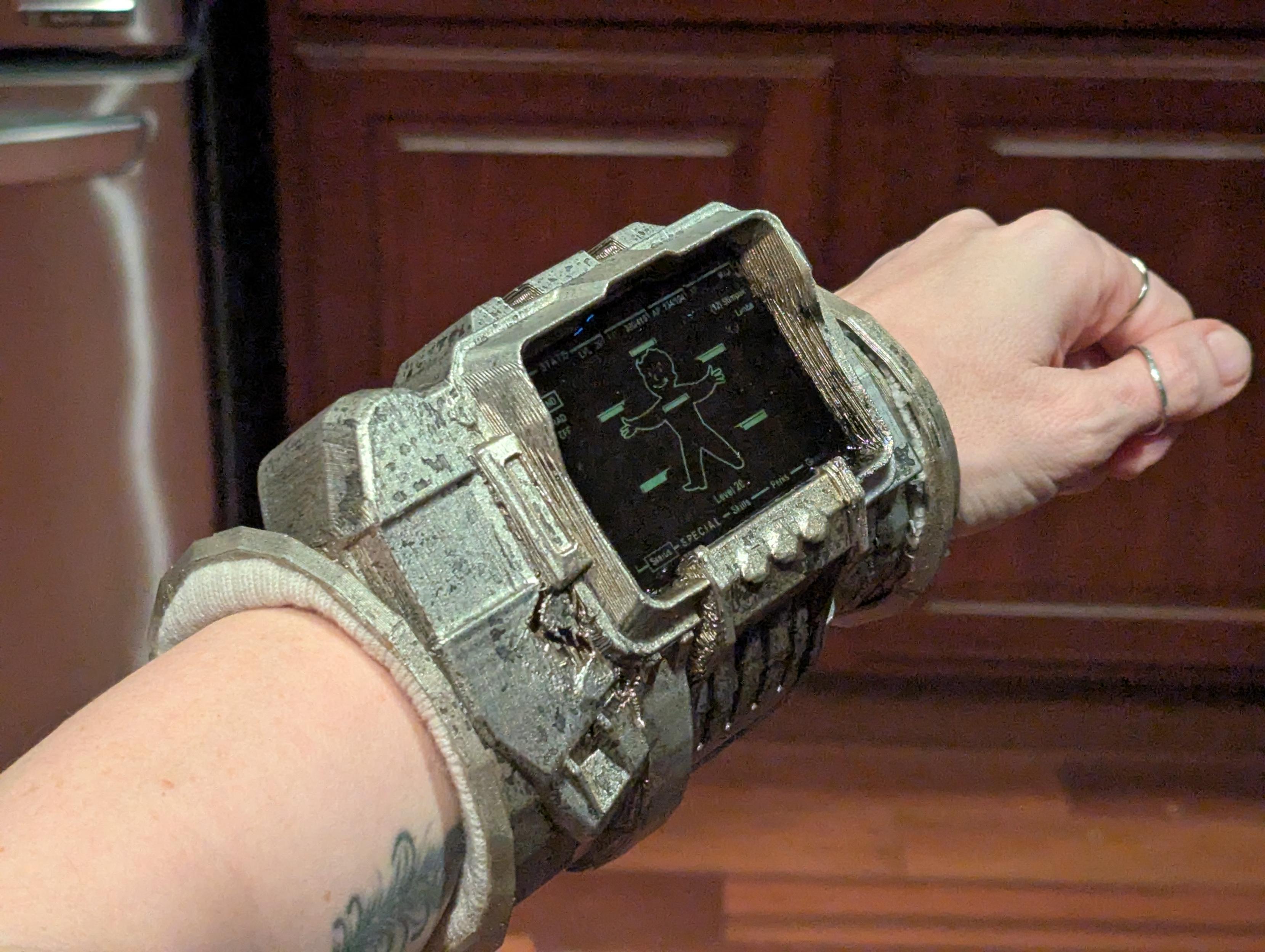 a costume pipboy on a woman's arm in front of dark wood cabinets and flooring. pipboy is an arm computer with a tiny CRT display, it's color is silver but not shiny, except for the screen