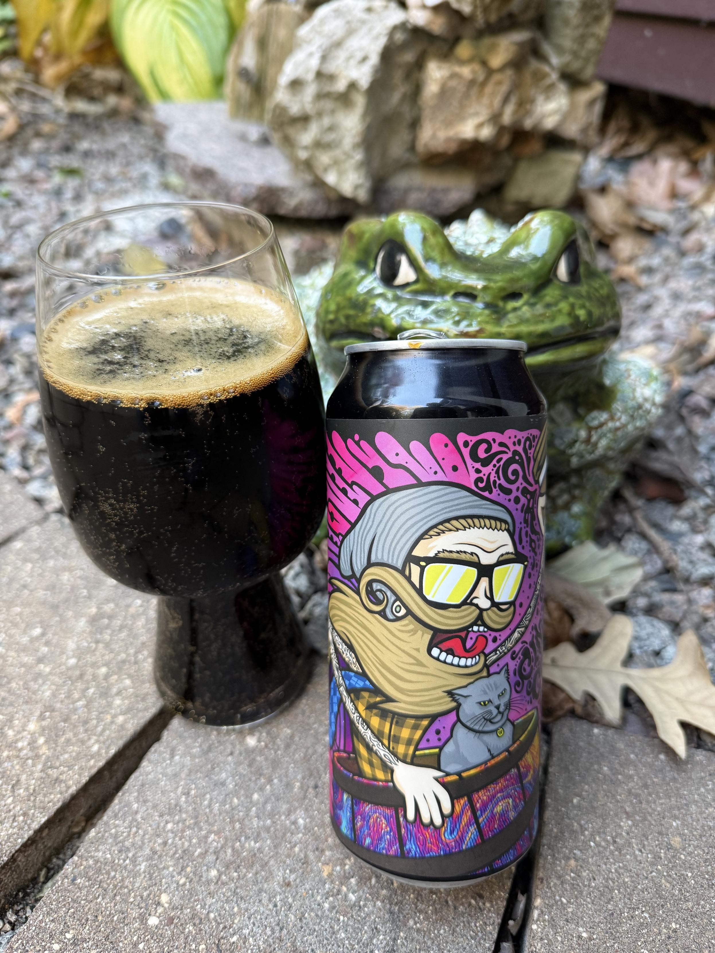 A stout glass of dark beer sits next to a pint can picturing a bearded man with a cat in his bike basket riding fast. In the background is a ceramic green toad