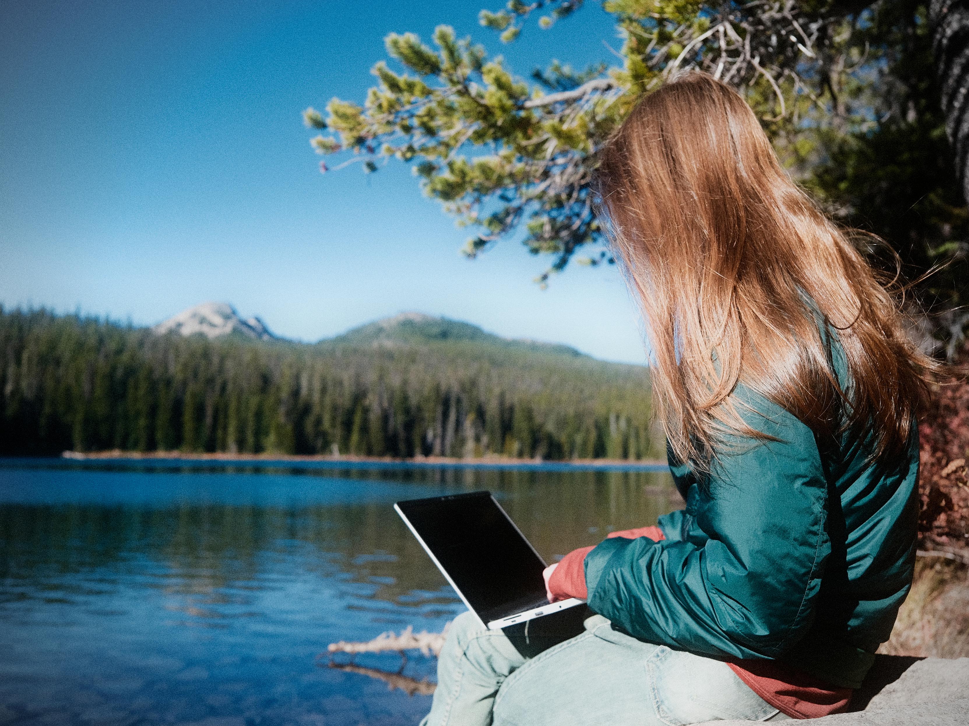 a woman on her laptop alongside an alpine lake with a forest of evergreen trees and mountains in the background