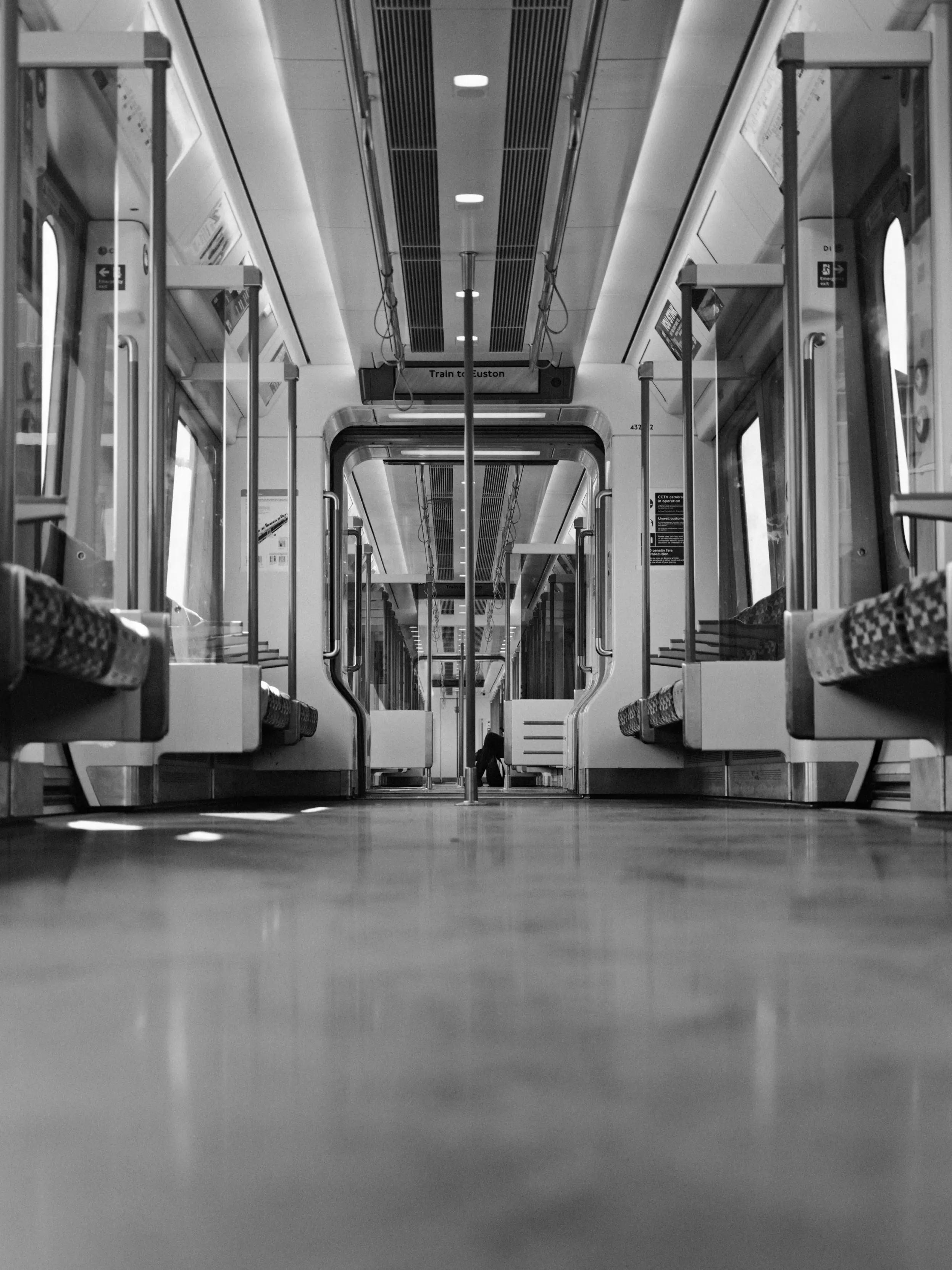 A black-and-white, low-angle perspective shot looking down the empty interior of a modern commuter train carriage. Polished grab poles run vertically and horizontally along rows of patterned, upholstered seats facing inward. Overhead vents, glowing rectangular lights, and reflective glass windows line the corridor, leading toward a distant doorway with a sign reading "Train to Euston".