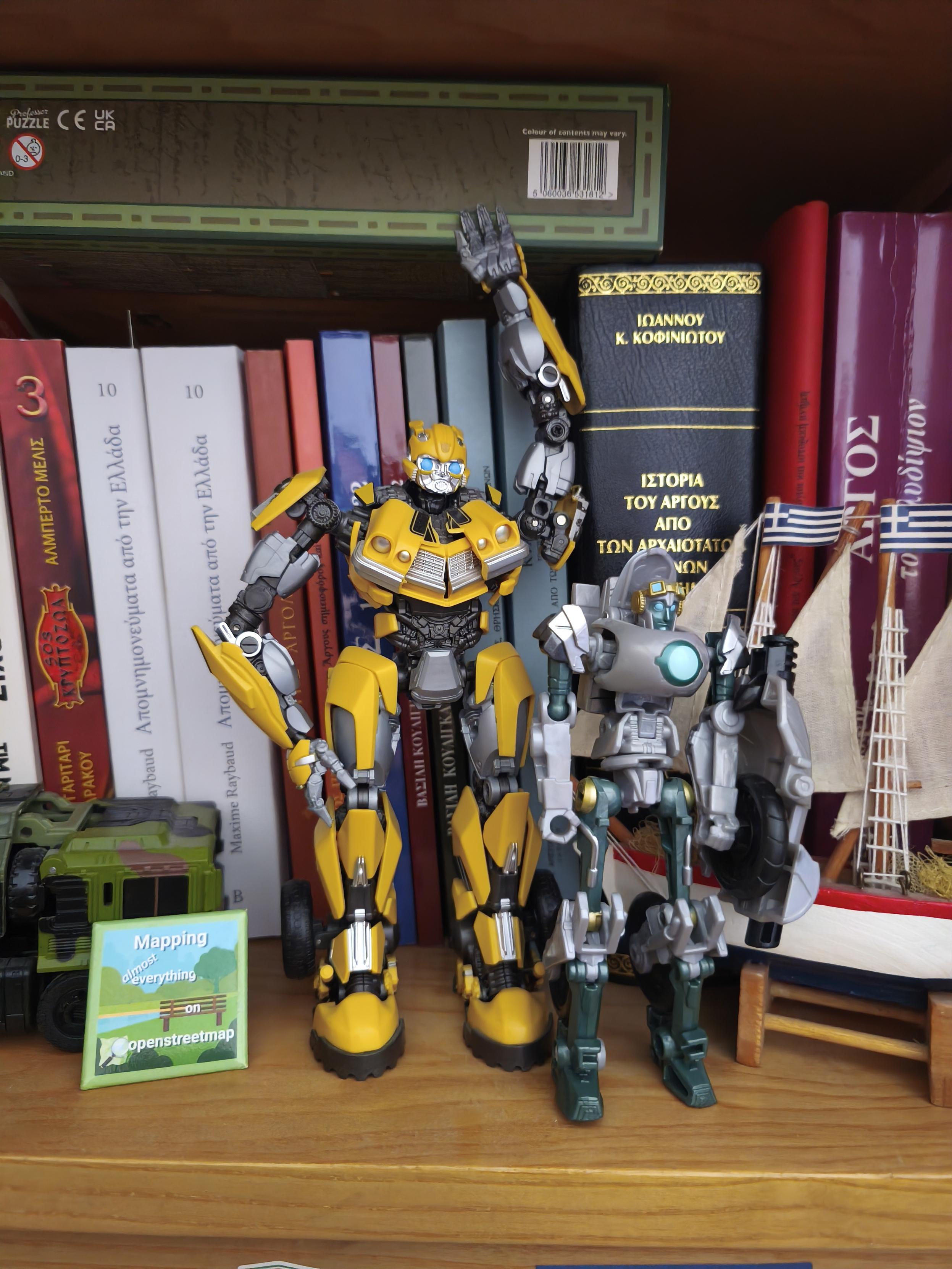 Photo from part of my office, with prominent the Transformers figures of Bumblebee (Rise Of The Beast) and Thrash (Earthspark), next to them an OpenStreetMap pin,on the other side a small boat with Greek flags, and behind all that stuff various books in Greek (some related to Argos and some other related to Nea Kios).