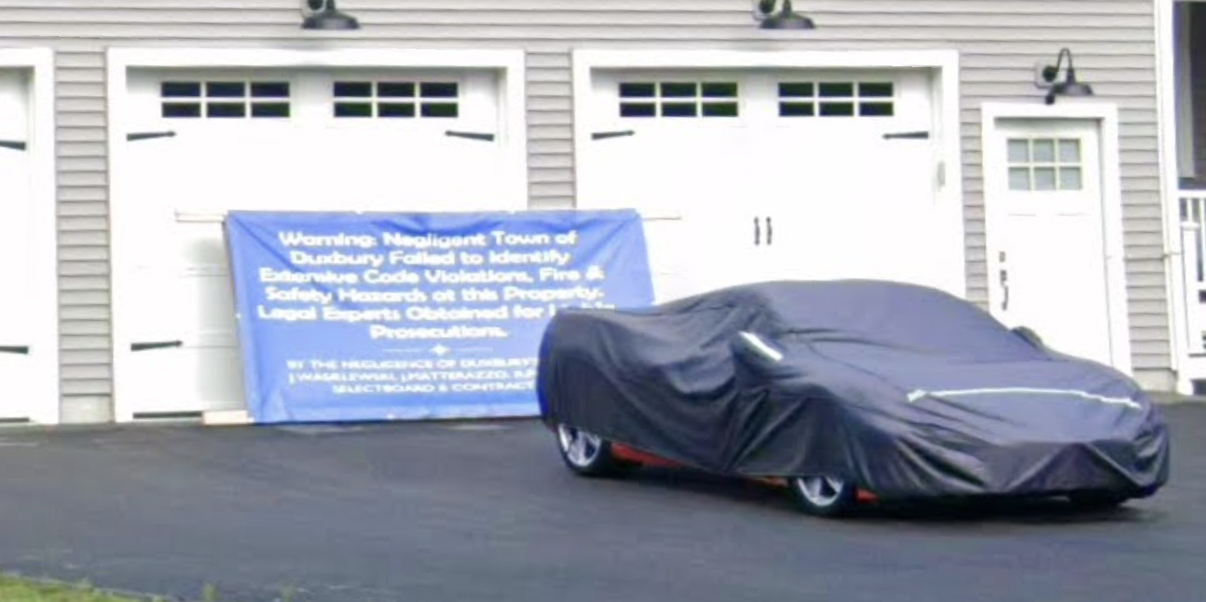 a zoom to show that there is a big angry sign stuck up against the garage: "WARNING: Negligent Town of Duxbury Failed to Identify Extensive Fire Code Violations, Fire and Safety Hazards on this property. Legal experts obtained for something-something prosecution"
in front of the sign is parked what I assume is a very expensive red sportscar in a protective cover, because they can't put it in the deathtrap garage.