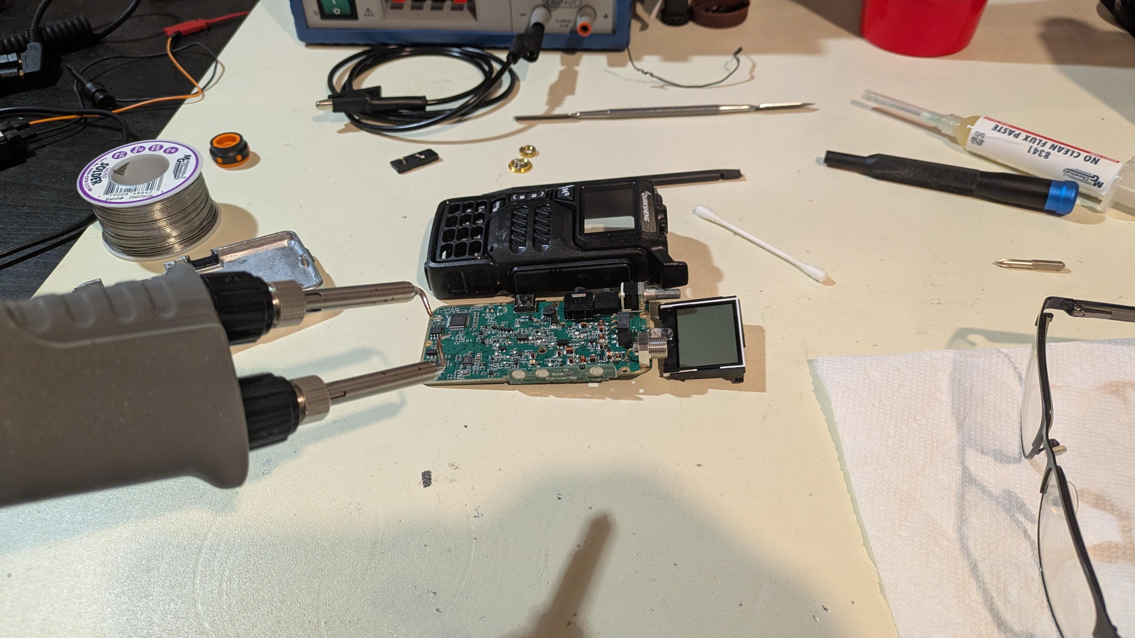 metal soldering tweezers in front of a messy desk with a disassembled handheld radio on it