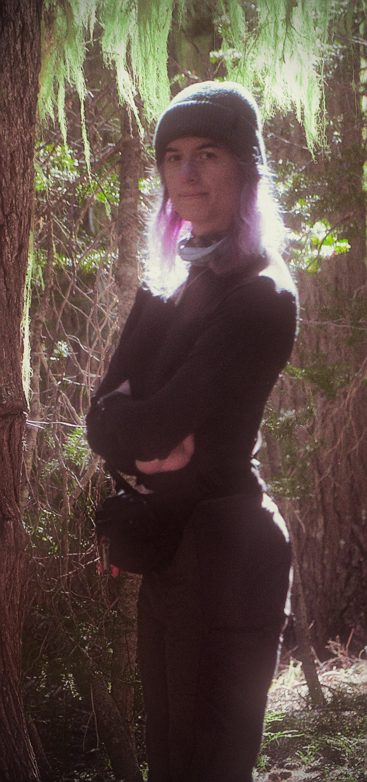 portrait of raven in a forest with the sun shining through behind her and green moss flowing off of trees behind her. her arms are crossed and she is wearing a green toque